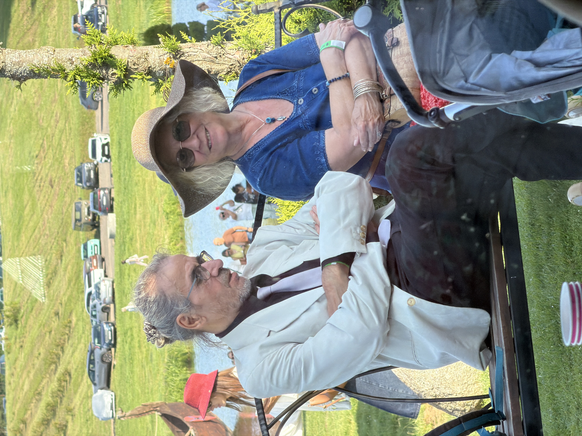 Two people are seated on a park bench outdoors. A man with grey hair pulled back in a bun and wearing glasses, a white suit jacket, and a light purple tie is on the left. He has his arms crossed. Next to him on the right is a woman wearing a wide-brimmed straw hat, sunglasses, and a blue dress. She is also wearing a necklace and bracelets, and her arms are crossed. The background shows a body of water, a grassy hill, and several parked cars. A large tree is to the right of the woman, and a stroller is partially visible in the foreground. The scene appears to be a public event or gathering, possibly in the area of Літвенскі сельскі Савет, Belarus, given the context of the provided information.