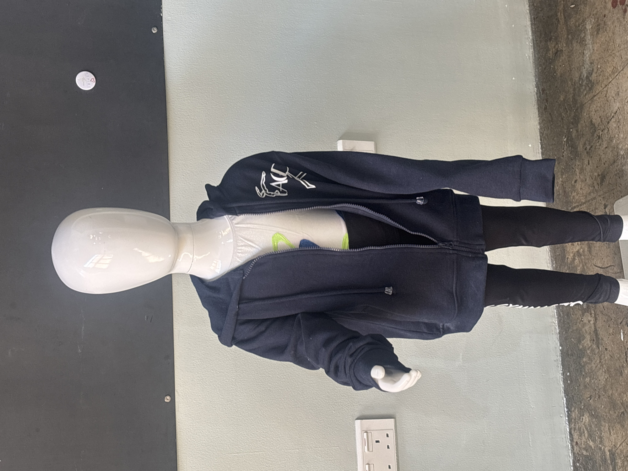 A full shot shows a white mannequin dressed in children's athletic wear. The mannequin wears a white t-shirt with a graphic design, a dark blue zip-up hooded jacket, and black leggings with white lettering down the side. The mannequin stands in front of a light blue wall and a dark gray wall. A white electrical outlet is visible on the left side of the blue wall. The floor is a textured gray concrete. The image was taken at the Dubai Mall, United Arab Emirates.