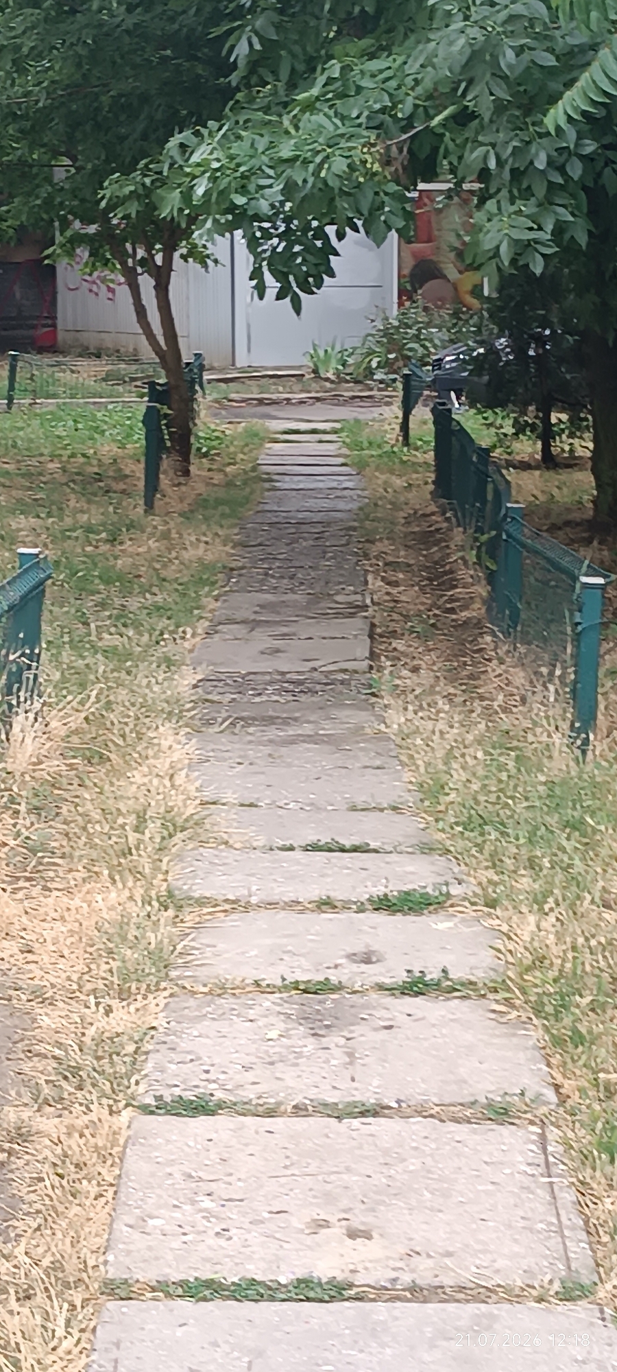 A narrow, overgrown pathway made of concrete slabs leads through a grassy area with sparse, dry vegetation. The pathway is bordered by a low, dark green metal fence on both sides. In the background, several trees with dense green foliage are visible, and beyond them, a light-colored building with a white door can be seen. The image appears to have been taken during the daytime, with natural light illuminating the scene. The timestamp "21.07.2026 12:18" is visible in the lower right corner of the image. The overall impression is one of an unkempt, outdoor space within a residential or park-like setting.