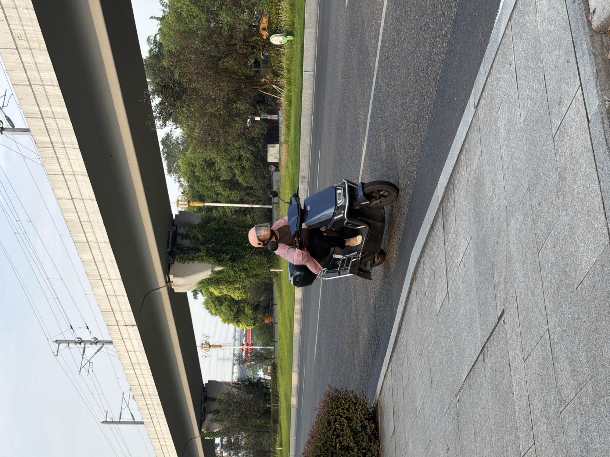 A person wearing a pink helmet with a clear face shield and a pink jacket over a brown top rides a dark blue electric scooter on an asphalt road in Jiangjin District, China. The scooter has a black storage box on the back. The person is looking forward and appears to be in motion. To the left of the road is a sidewalk made of grey paving stones. To the right of the road, there is a grassy area with trees and bushes. In the background, a large elevated concrete structure, likely a bridge or overpass for a railway or highway, spans across the scene. Overhead power lines are visible on the structure. The sky is light blue with some wispy clouds, suggesting it is daytime. The lighting indicates it is likely late afternoon or early morning due to the soft light.