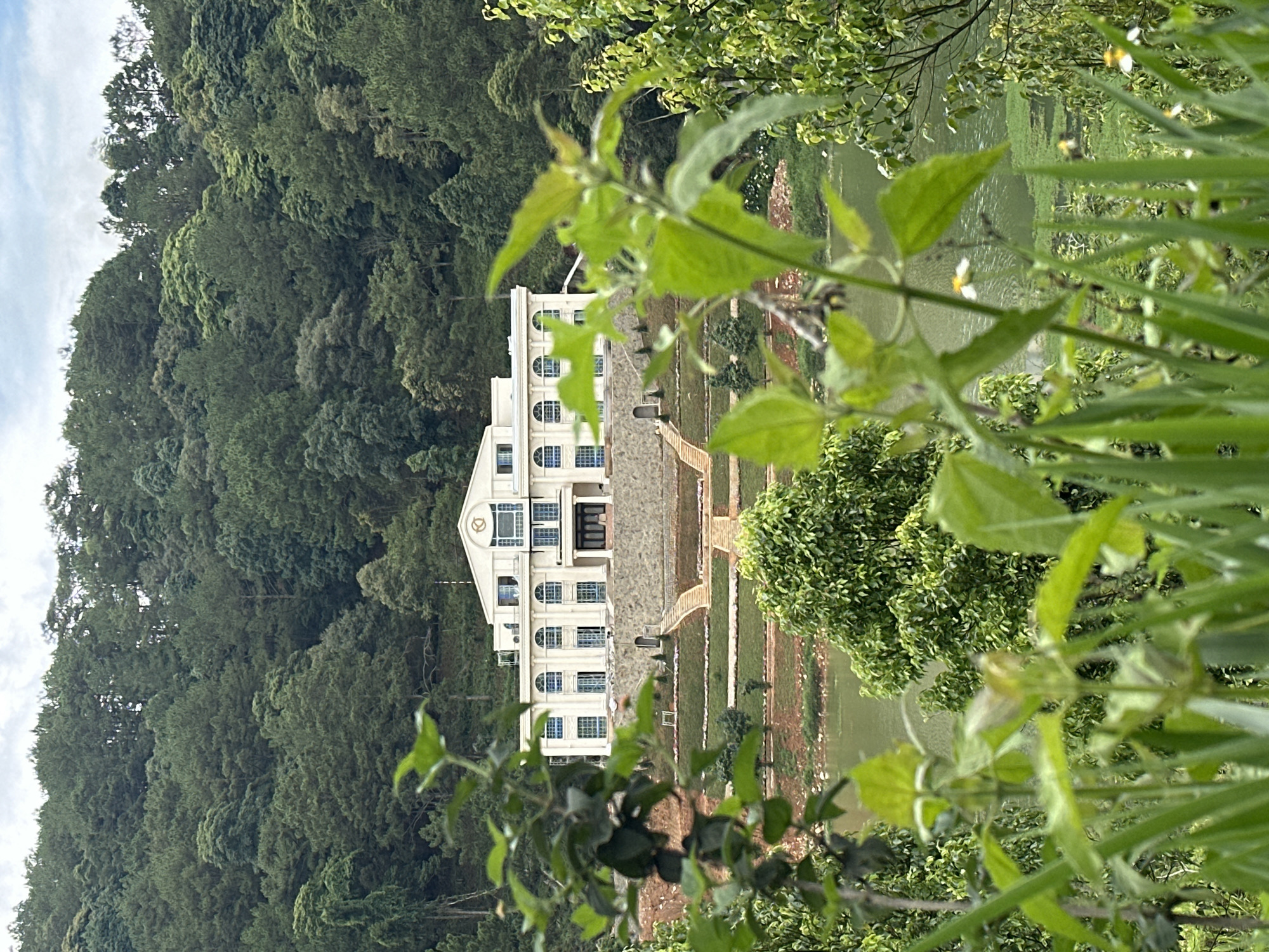 The photograph depicts a large, multi-story white building situated in Da Lat, Vietnam. The building has a symmetrical facade with arched windows and a prominent clock tower-like structure at its center. It is surrounded by a well-manicured lawn that slopes down to a body of water. Dense green foliage, likely part of a forest or hillside, borders the property. The foreground is partially obscured by foliage, creating a framed view of the building and its surroundings. The sky is partially visible with some cloud cover.