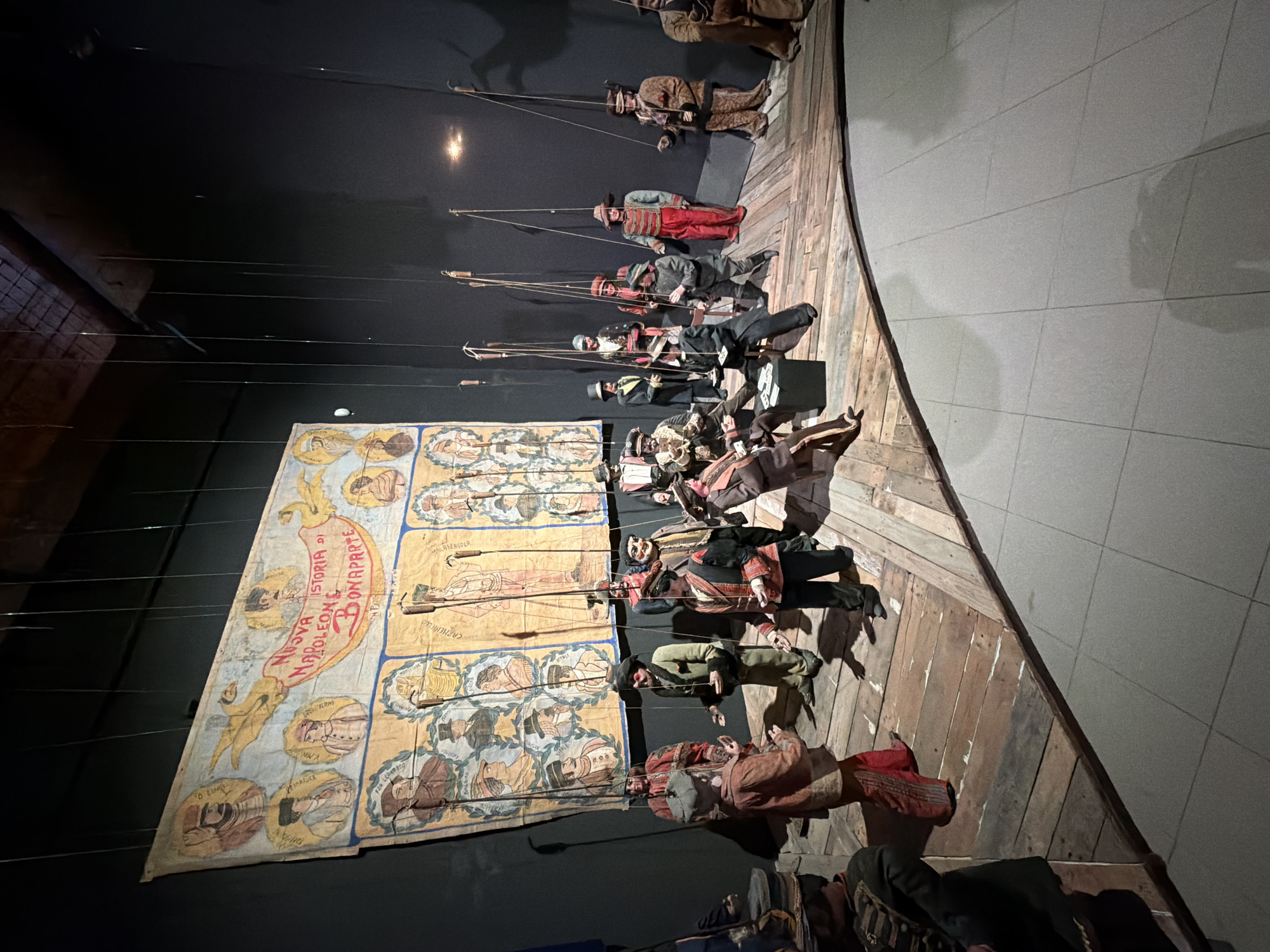 This image depicts a puppet show display, likely part of a museum exhibit, set in Palermo, Italy. The scene is centered around a large, hand-painted backdrop featuring portraits and the text "NUOVA STORIA DI NAPOLEONE BONAPARTE" (New History of Napoleon Bonaparte). Below the backdrop, a collection of marionettes are arranged on a wooden stage, some standing and others seated. The puppets are dressed in historical costumes, representing various characters. One group of puppets appears to be engaged in a card game. The stage is constructed from aged wooden planks and is elevated from a tiled floor. The overall environment is dimly lit, suggesting an indoor theatrical setting or museum display. The puppets are suspended by strings, indicating they are operated from above.