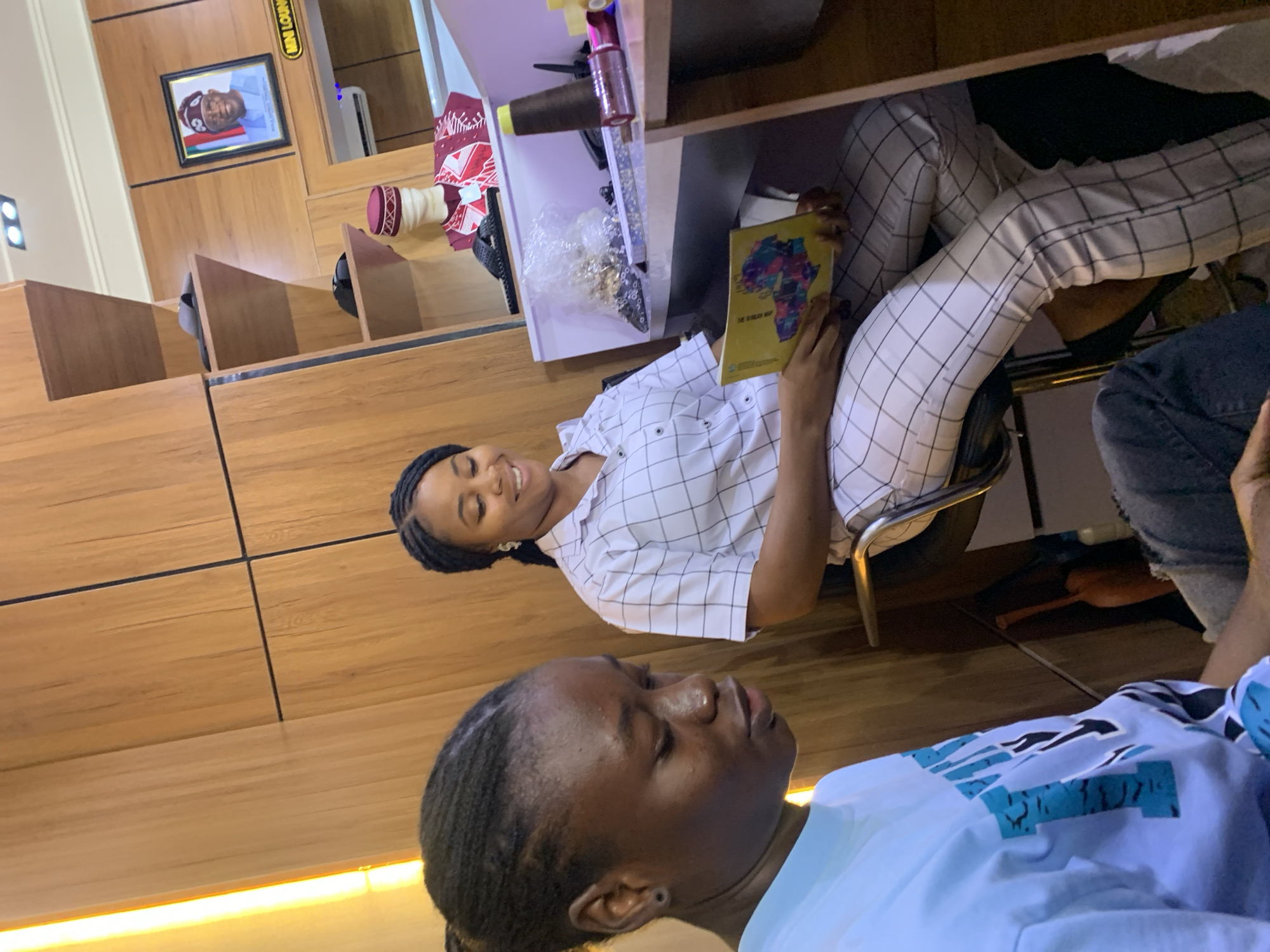 The image captures an indoor scene with two young women. The woman on the right, seated on a stool, is wearing a white collared shirt with a black grid pattern and matching pants. She is holding a yellow book open in her lap and smiling. The woman on the left is partially visible in the foreground, wearing a light blue t-shirt with a blue and black graphic design. She is looking down and away from the camera. The environment appears to be a retail or service establishment, possibly a tailor's shop or a boutique, given the presence of a mannequin displaying traditional clothing, spools of thread, and a framed portrait of a man in traditional attire on the wall. The wall behind the women is made of wood panels. The lighting suggests an indoor setting, possibly during the day. The text "MINI LOUNGE" is visible on a sign in the background, possibly indicating the establishment's name or a section within it. The overall impression is a casual and possibly relaxed atmosphere. There is no explicit visual information to pinpoint the location to Gura Top, Nigeria.