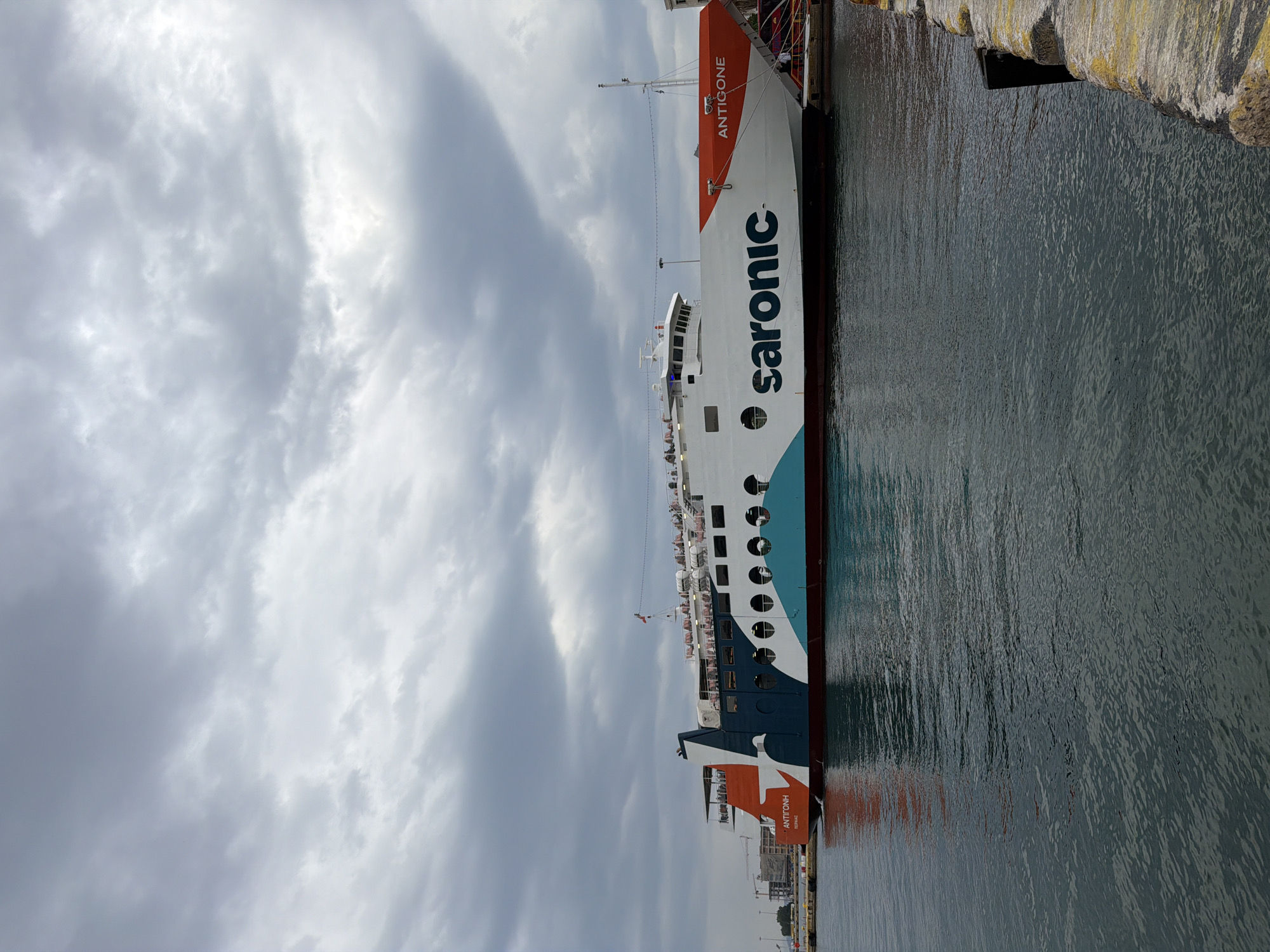 A ferry boat, named "Antigone" of the "Saronic Ferries" line, is docked in the harbor of Piraeus, Greece. The boat is white with orange and blue accents, featuring the company's name in large, bold letters. The sky overhead is overcast with heavy, grey clouds. The water in the harbor is calm and reflects the colors of the boat and sky. A stone pier is visible beside the boat.