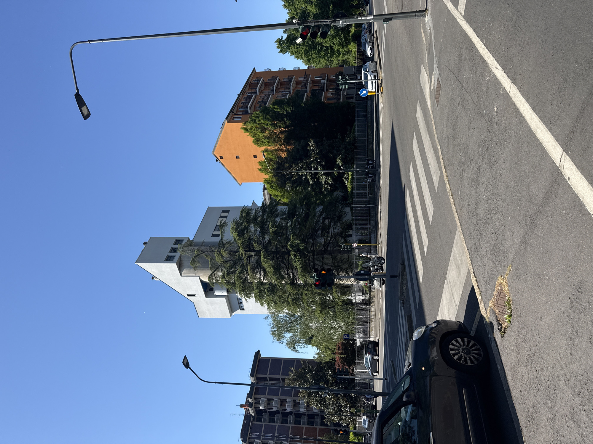 The image depicts a street scene in Milan, Italy, under a clear blue sky. The foreground shows a portion of a black car parked on the side of the road, near a pedestrian crosswalk marked with white stripes. The street surface is asphalt.
In the middle ground, there are multiple buildings of varying architectural styles and colors. A white, modern-style building stands out, along with a brown brick building and an orange residential building. Trees are interspersed among the buildings, partially obscuring the facades.
Traffic lights are visible on the street. The color of the traffic lights are red. A directional street sign indicates the direction of travel for vehicles. Motorbikes are also parked in the vicinity.