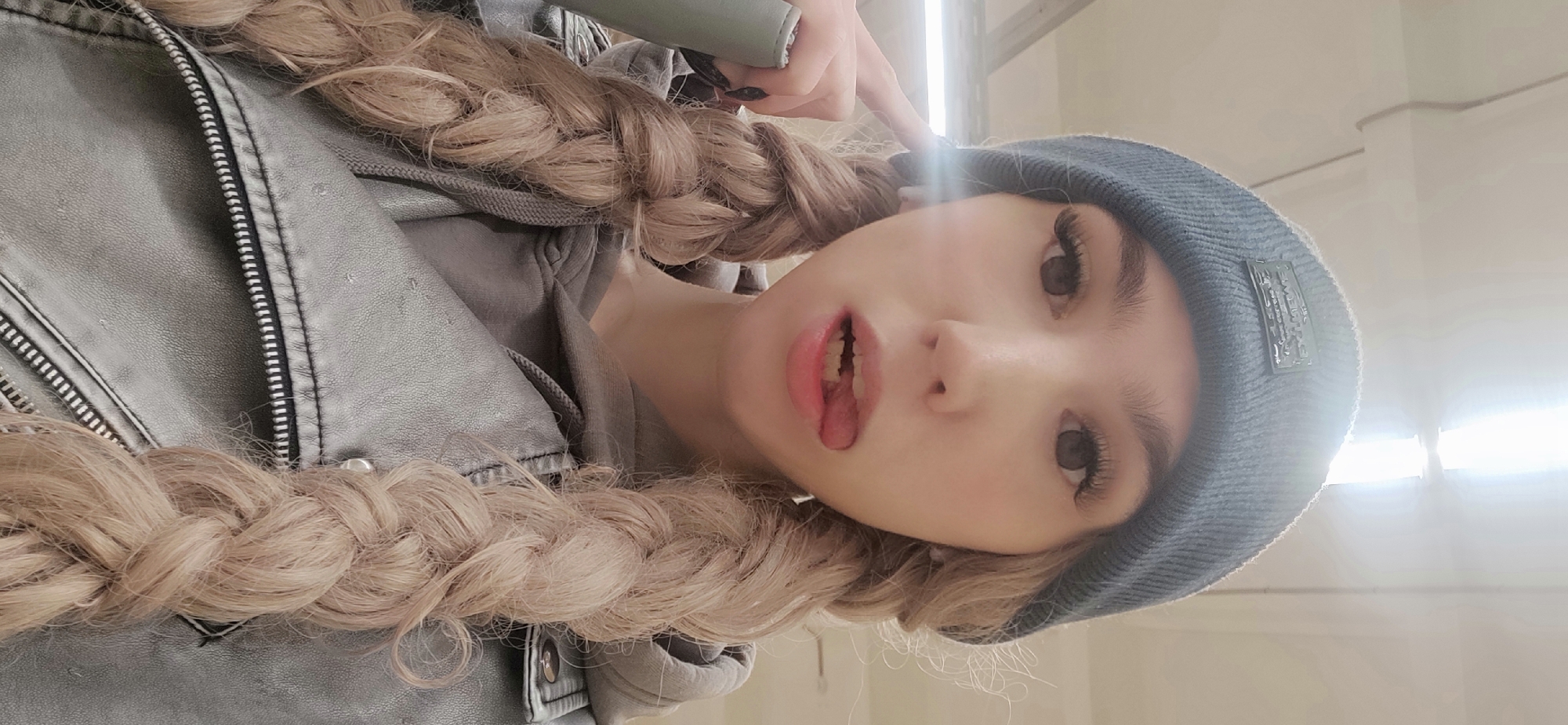 A young woman is posing for a selfie indoors. She is wearing a gray beanie, a gray jacket with a zipper, and has blonde hair styled in two braids. The location is the city of Chaikovsky, Russia.