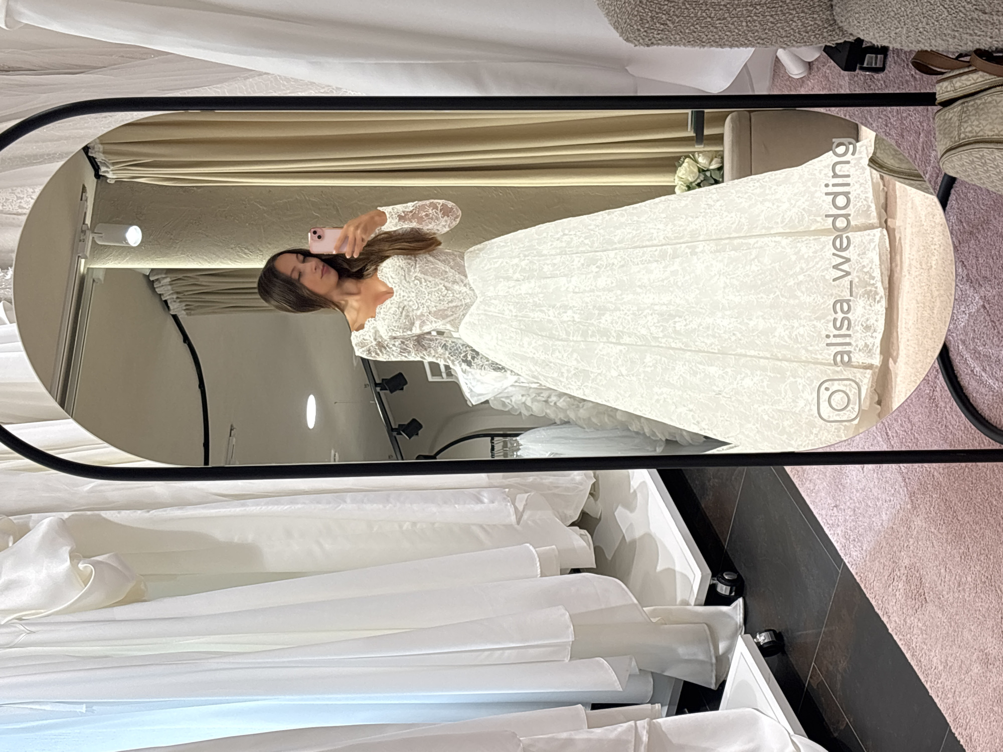 A mirror in a bridal shop in Saint Petersburg, Russia, reflects a woman taking a selfie in a white lace wedding dress. The mirror is oval, framed in black metal, and shows the woman holding a pink phone, the dress, and a glimpse of the shop's interior, including a rack of white gowns. The background reveals elements of the fitting room: beige curtains, decorative flowers, and lighting fixtures. The woman's reflection is partly obscured by the Instagram handle "@alisa_wedding." Outside the mirror, the flooring alternates between black and pink and a partially visible sofa. Below the mirror, a collection of white dresses is visible in a container.