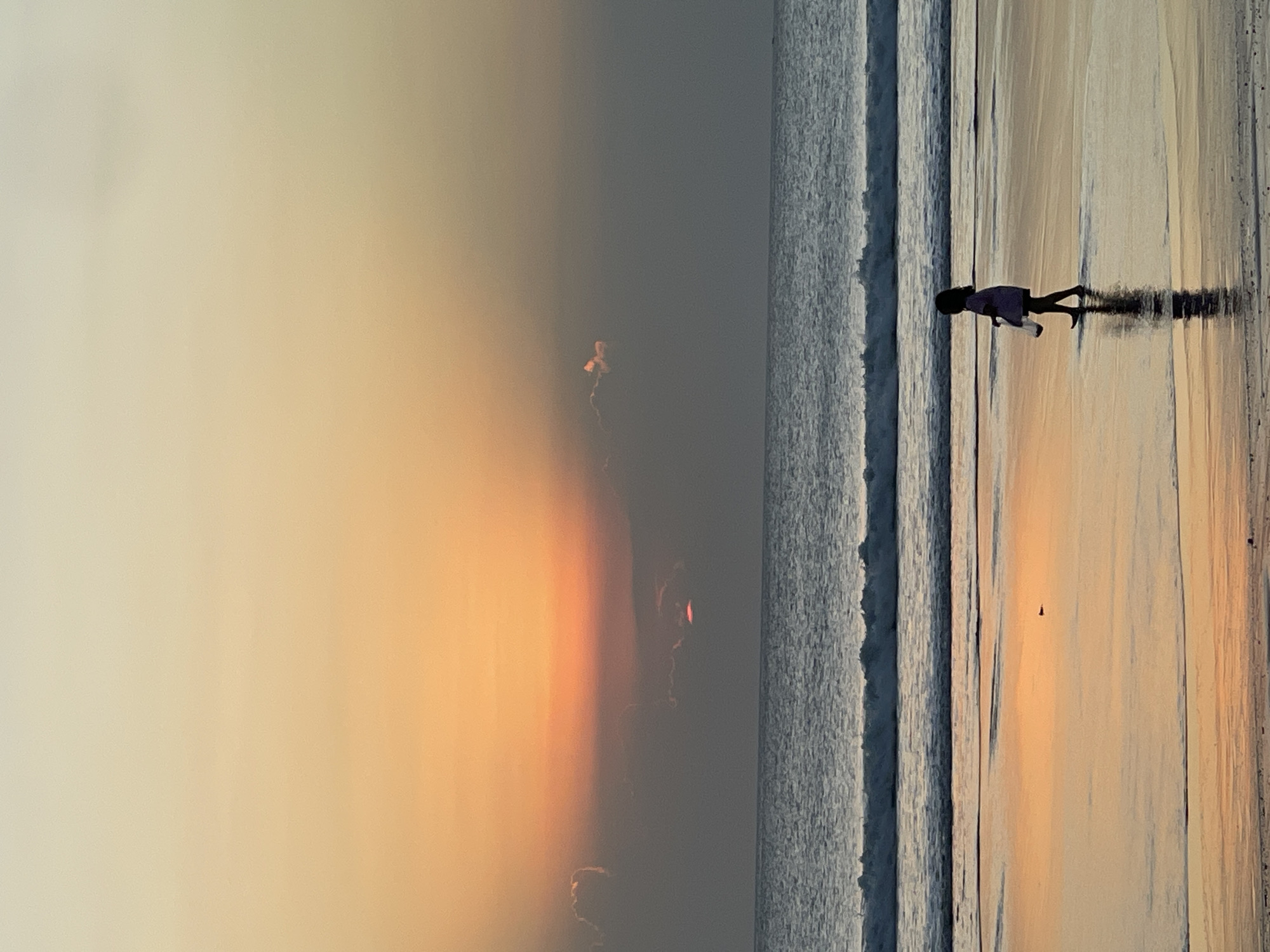 A young child walks on the wet sand of Kuta Beach in Bali, Indonesia, during sunset. The child, silhouetted against the shimmering sand and water, is wearing a light-colored dress and carrying an object in their left hand. The ocean stretches out to the horizon, with gentle waves rolling onto the shore. The sky is a canvas of soft orange and yellow hues, with the sun partially obscured by clouds. The reflection of the sunset is visible on the wet sand, creating a serene and picturesque scene.