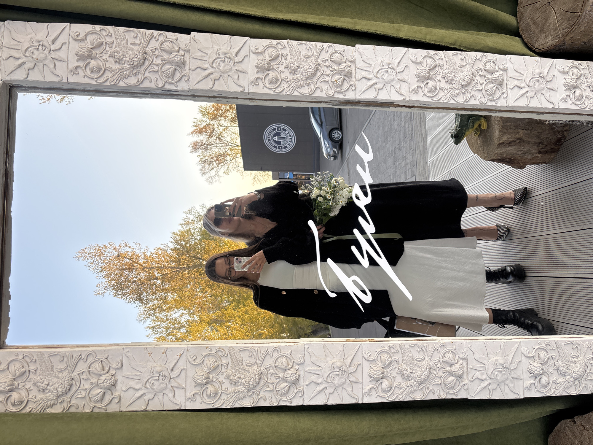 Two women are reflected in a decorative mirror, holding phones as if taking a selfie. One woman wears a white dress with a black blazer and black boots, while the other wears an all-black outfit with patterned shoes. One holds a small white bouquet of flowers. In the background of the mirror's reflection, there is a dark building with a round logo and a car parked nearby. The mirror has an ornate, white frame with floral and sun motifs. The mirror appears to be placed outdoors, possibly on a patio with wooden decking. According to the metadata the location is Krasnoyarsk, Russia.
