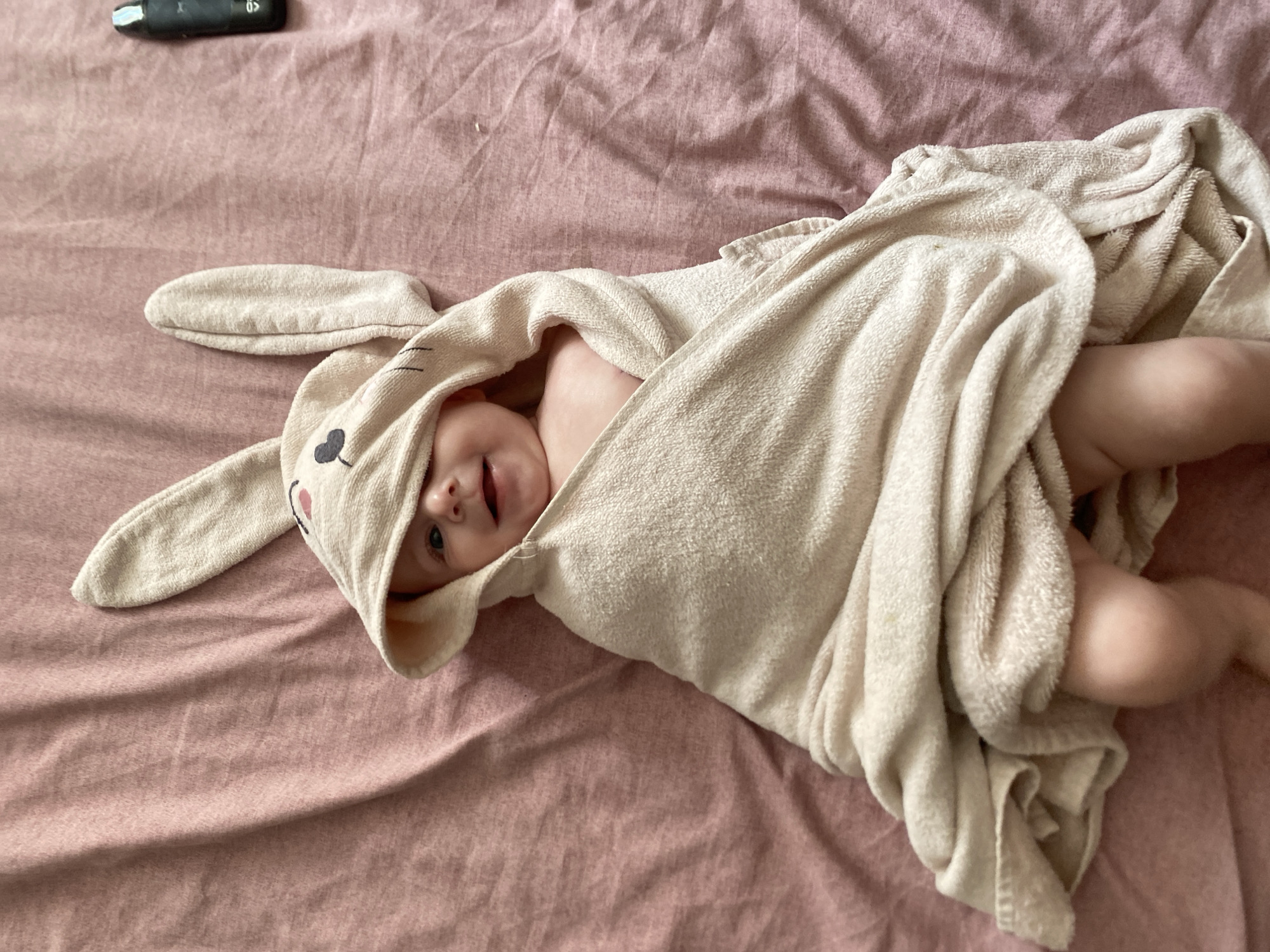 A baby wrapped in a beige towel with bunny ears is lying on a pink blanket. The baby is smiling and looking at the camera. There is a black object on the right side of the image. The image appears to be taken indoors.