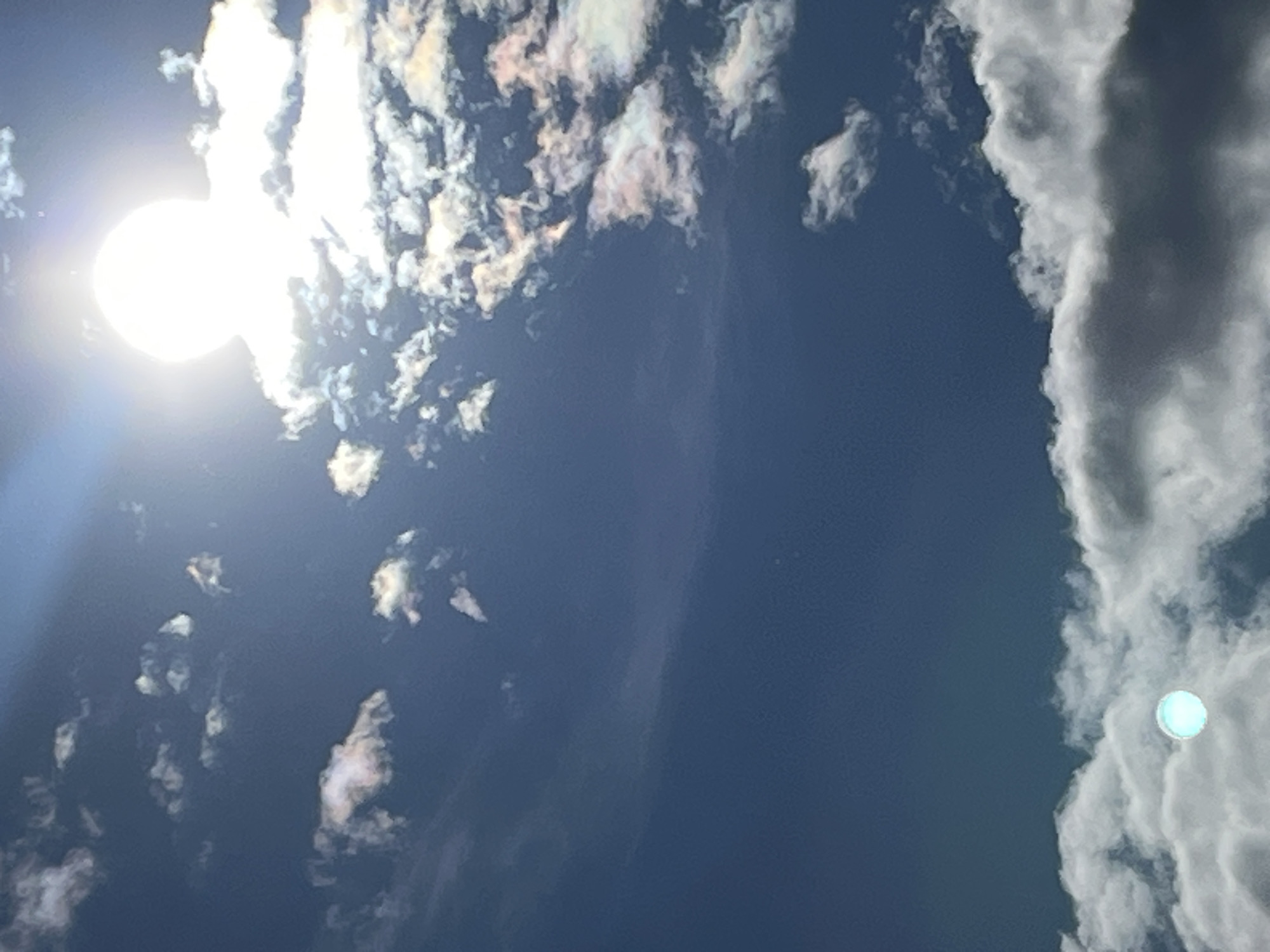 The image shows a daytime sky in Chambœuf, France, with a bright sun obscured by thin clouds in the upper left corner. Smaller, scattered clouds are visible throughout the sky. A dense band of clouds is present on the right side of the frame. The sky itself appears to be a deep blue.