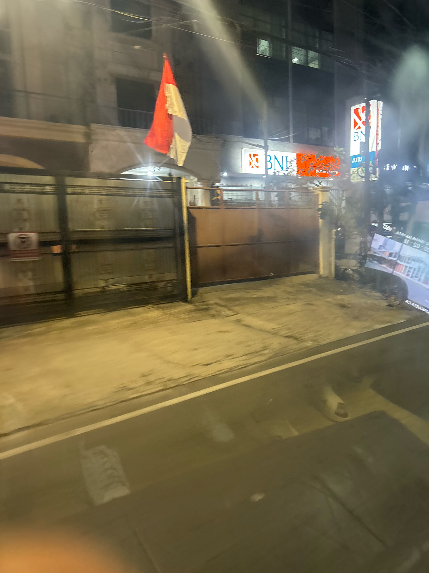The image captures a street scene in North Jakarta, Indonesia at night. In the foreground, a street with a white dividing line is visible, reflecting the ambient light. The middle ground features a metal gate and a brown gate, with an Indonesian flag hanging from a wire above. Adjacent to the gates, a building with signage for "BNI" and "ATM" is illuminated. The building appears to be a bank. The background shows a multi-story building with darkened windows. The overall atmosphere is one of a quiet urban night, with elements of local culture and commerce present.