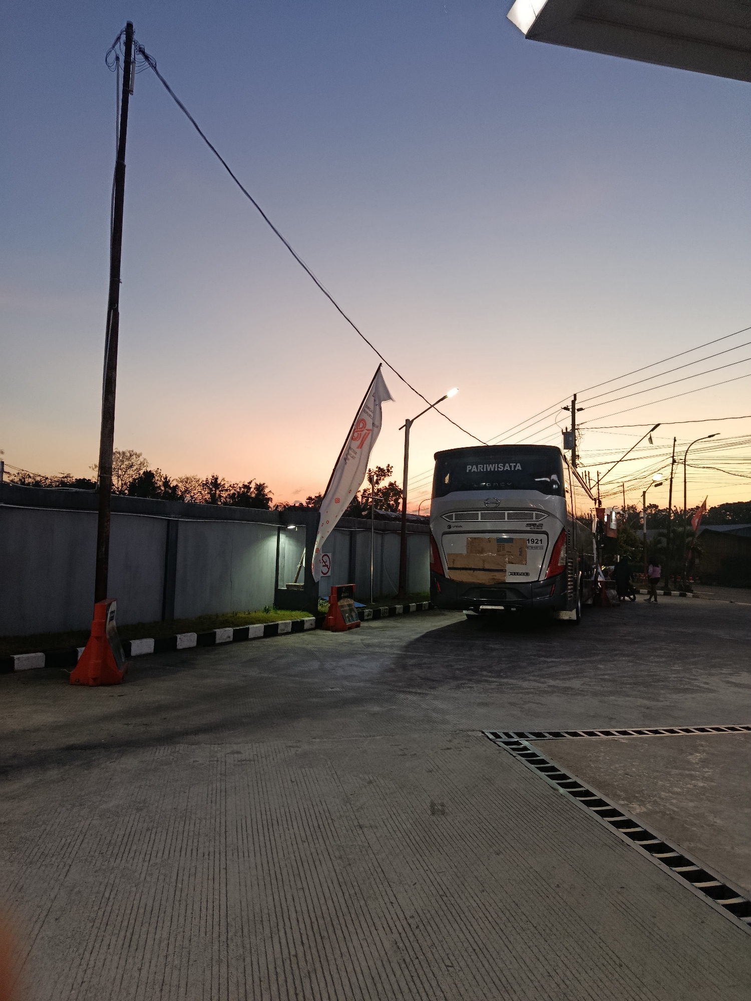 The image captures a view of a parking lot at dusk in Sedayu, Indonesia. A large tour bus, marked with "PARIWISATA" (tourism), is parked in the center. The bus is gray and white with a license plate displaying "1921". In front of the bus, a white flag with red lettering is displayed on a pole.
The surrounding environment features a concrete ground with striped patterns, a gray concrete wall along the left side of the frame, and several utility poles with electrical wires crisscrossing the sky. Orange traffic cones are visible near the wall. The sky displays a gradient of colors from deep blue at the top to orange and pink near the horizon, indicating sunset. In the background, trees and the outlines of buildings can be seen.
Two individuals are partially visible near the rear of the bus, appearing to be standing. The lighting suggests it is either early morning or late evening. The overall scene is an outdoor public space with signs of recent activity related to transportation.