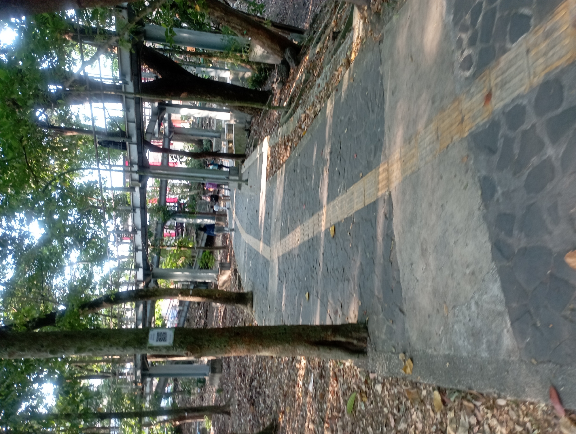 The image depicts an outdoor scene in Depok, Indonesia, featuring a paved walkway with textured tactile paving for accessibility. The walkway winds through a park-like setting with numerous trees, some of which have prominent trunks. In the background, a raised pedestrian bridge structure, likely made of metal and concrete, is visible, with several people walking on it. The ground is covered with fallen leaves, indicating it may be autumn or a similar season. The lighting suggests it is daytime, with sunlight filtering through the tree canopy, casting shadows on the walkway. The overall environment appears to be a public space designed for walking and recreation.