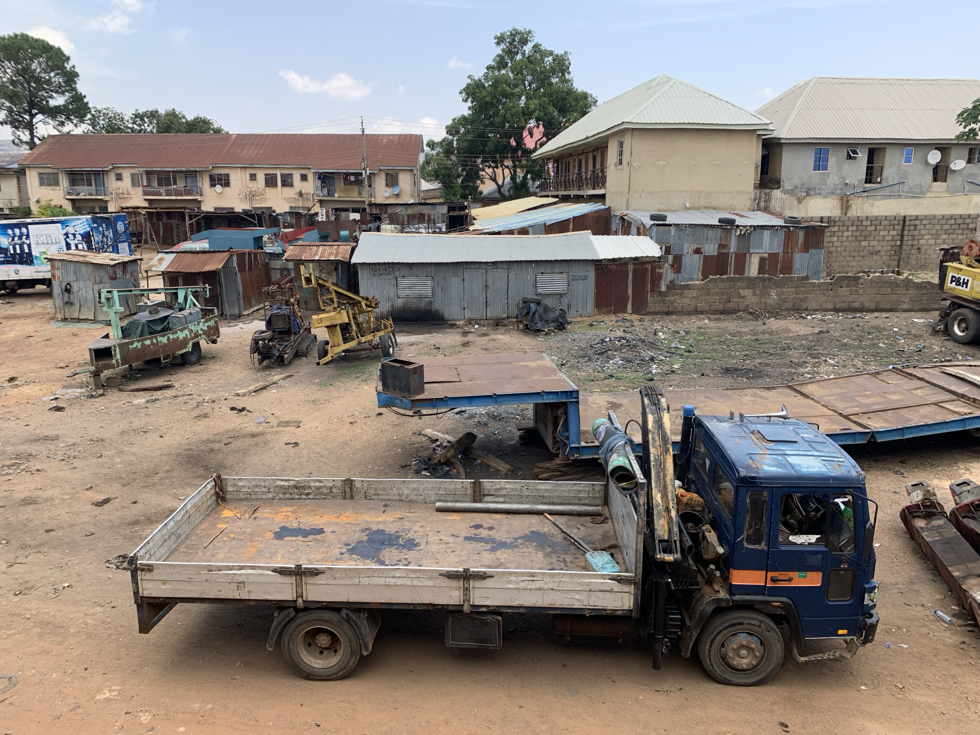 The image depicts a cluttered scene in Jos, Nigeria. A blue flatbed truck with a crane attached is parked in a dirt lot. The truck's bed is dirty, and various objects are scattered inside. In the background, there are makeshift corrugated metal structures, older brick buildings, and vegetation. Several other vehicles, including a yellow crane and what appears to be a rusted trailer, are visible in the lot. The sky is overcast. There are no people visible.