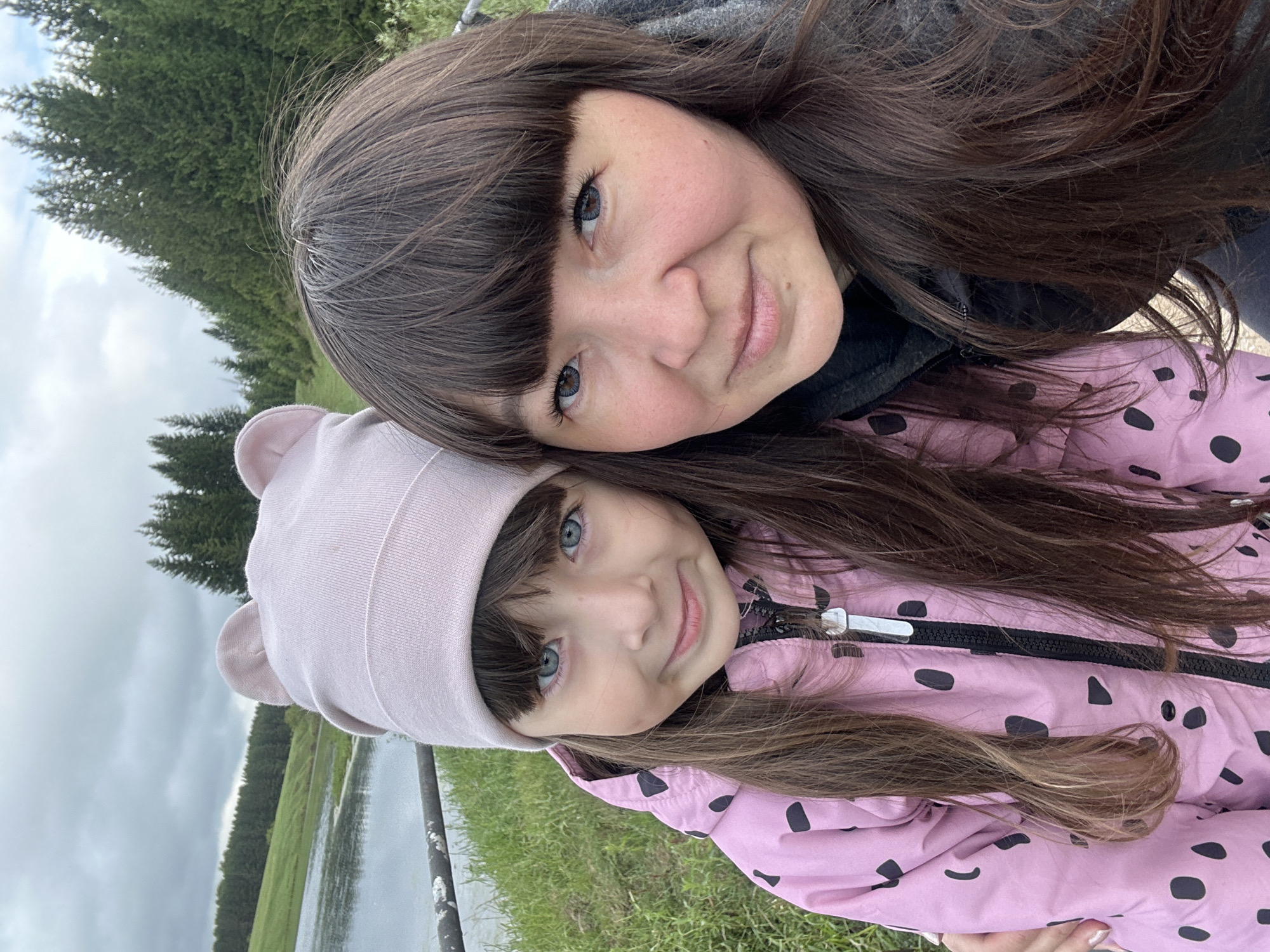 Two females are visible in this selfie. The older female has dark brown hair with bangs and appears to be wearing a dark gray hoodie. The younger female, presumably a child, has light brown hair, blue eyes, and is wearing a light pink beanie with ears and a pink jacket with black spots. They are positioned close to each other and smiling slightly, suggesting a close relationship. The background includes green grass, a body of water, trees, and a cloudy sky. The location appears to be Среднее Кечево, Russia, based on geotagging or other metadata, as the setting implies a nature area near a village or town.