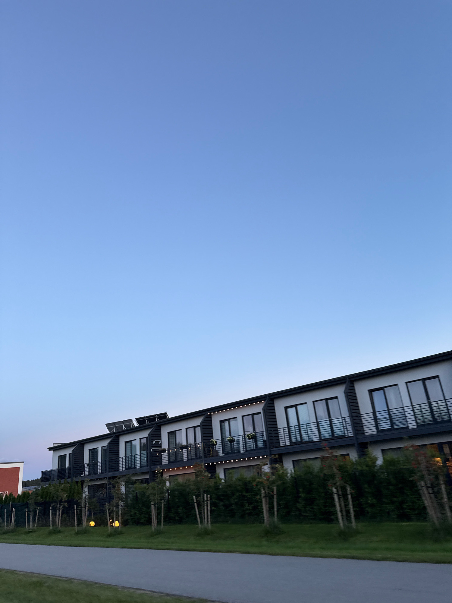 The image shows a row of modern townhouses in the city of Alliku, Estonia. The houses are two stories tall with white facades and dark roofs. Each unit has large windows and balconies with black railings. Several units have solar panels installed on their roofs. In front of the townhouses, there is a neatly trimmed green hedge and several young trees planted in a row. A paved road runs in front of the property, with a grassy area between the road and the hedge. The sky is clear and a pale blue, suggesting it is either early morning or late evening. There are no people visible in the image.