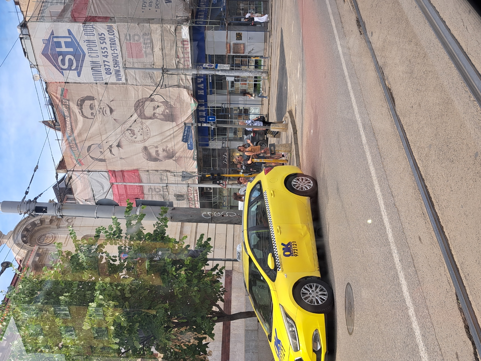 The image captures a street scene in Sofia, Bulgaria, on a sunny day. A bright yellow taxi, clearly marked with "OK TAXI 972121," is parked alongside tram tracks. In the background, a building is covered in large banners, one featuring the logo "SH" and the Bulgarian text "ШПИЦИ СТРОЙ ООДУ" along with a phone number and website. Another banner displays portraits of historical figures, identified by the text "NATIONAL HEROES." Several people are visible, some standing near the taxi and others walking in the distance. The presence of scaffolding suggests ongoing construction or renovation work on the building. The overall atmosphere appears to be that of a bustling urban environment.