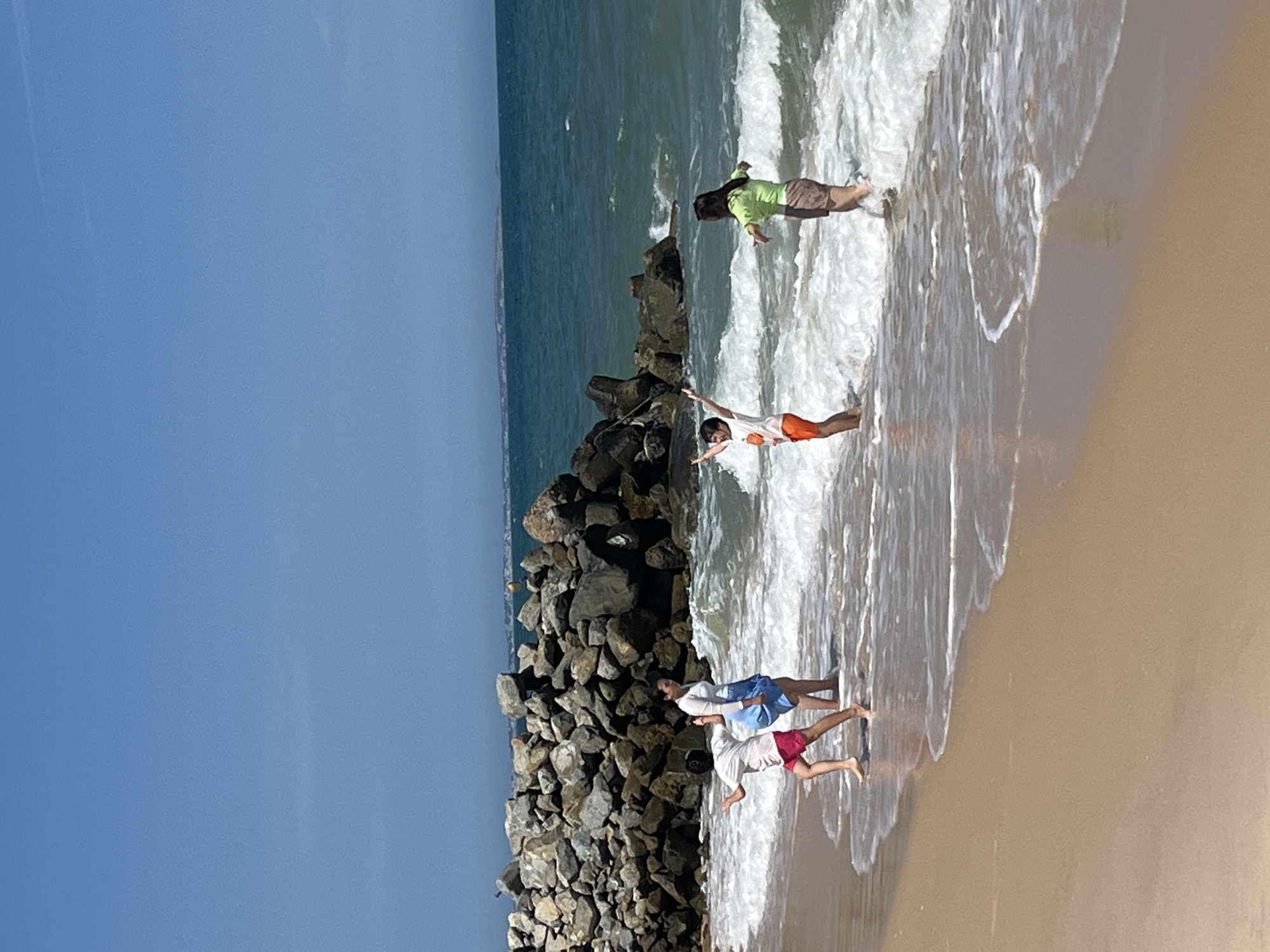 In Mũi Né, Vietnam, four individuals are visible on a sandy beach near a rocky breakwater. The ocean's waves gently lap at the shore, creating a foamy surf. The sky above is a clear, bright blue.
On the left, a person wearing a white t-shirt and red shorts stands with their back to the viewer, appearing to be running or moving quickly. To their right, another individual in a white top and blue skirt is standing, facing away from the camera.
In the middle, a person with their arms raised in the air, wearing an orange t-shirt and orange shorts, is captured mid-action. To their right, a person in a light green shirt and brown shorts stands with their arms spread wide.
The scene suggests a recreational activity, possibly playing in the water or posing for a photograph. The environment is natural, with the beach, sea, and rocks creating a scenic backdrop. The focus is on the individuals and their playful engagement with the ocean.