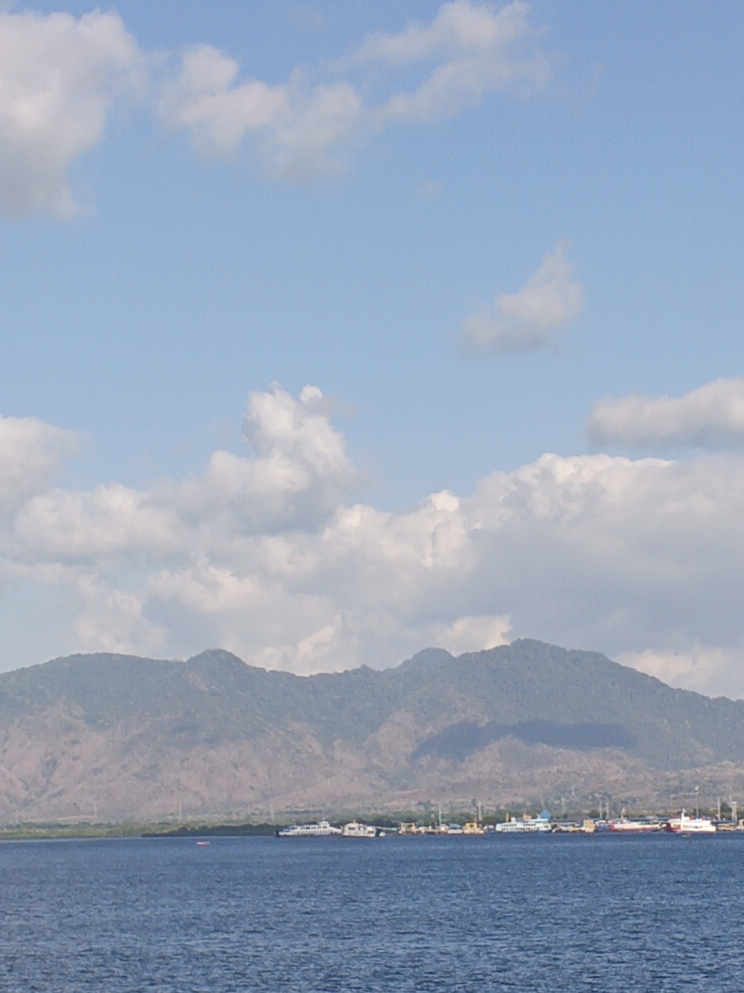A wide shot captures a calm, blue ocean in the foreground with a distant shoreline and a mountain range under a partly cloudy sky. The shoreline features several boats docked, including ferries and smaller vessels, suggesting a port or harbor. The mountains in the background are a muted green and brown, with visible textures and shadows indicating rugged terrain. White, puffy clouds dot the sky, contrasting with the pale blue expanse. The scene evokes a sense of a tranquil coastal environment in Indonesia.