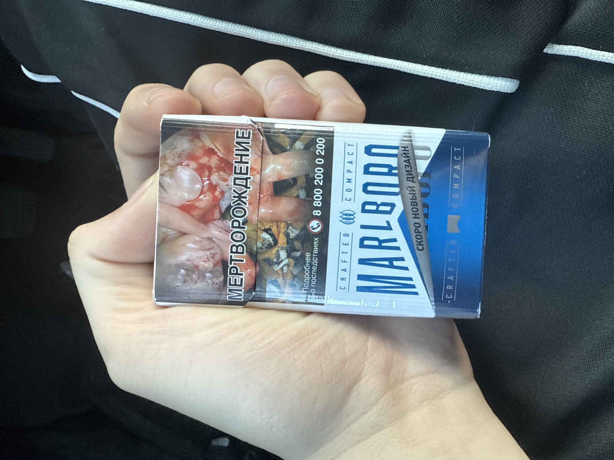 A person's hand is holding a pack of Marlboro cigarettes. The pack has a warning label depicting a graphic image of what appears to be damaged internal organs and the text "МЕРТВОРОЖДЕНИЕ" which translates to "Stillbirth" or "Abortion." Below this, there is text in Russian and a phone number. The brand name "MARLBORO" is prominent on the pack, along with "COMPACT" and "СКОРО НОВЫЙ ДИЗАЙН" which translates to "New Design Soon." The background is blurred and appears to be the interior of a vehicle. There is no identifiable information to suggest the location is Samara, Russia.