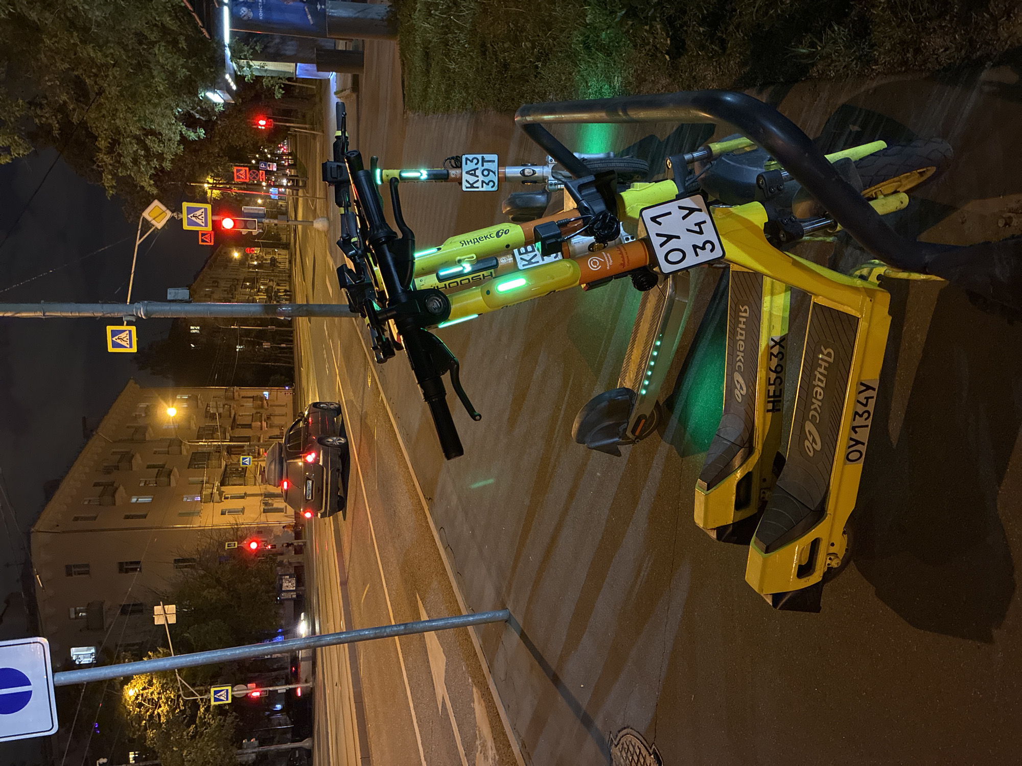 The image shows two shared electric scooters parked on the side of a road at night in Moscow, Russia. The scooters are yellow and black, with visible license plates: "OY134Y" and "KA339T." Logos for "HOOSH" and "Яндекс Go" are displayed on the scooters. The scooters are parked adjacent to a sidewalk, with a street lamp and various road signs visible nearby, including pedestrian crossing signs. Streetlights and traffic lights are visible in the background along with buildings and a car with illuminated taillights. The environment appears to be a city street at night.