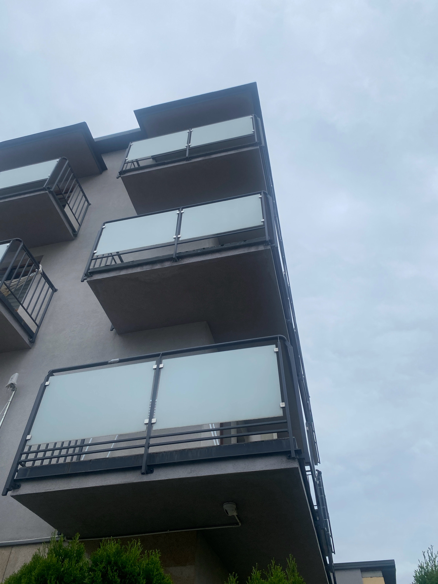 The image shows a close-up, low-angle view of a modern apartment building with multiple balconies, likely in Nowy Sącz, Poland. The building's exterior is a muted gray, and the balconies feature sleek, glass-paneled railings supported by dark metal frames. The sky above is overcast and a uniform light gray, contributing to a somewhat muted and diffused lighting condition. Some green foliage, possibly bushes or small trees, is visible at the bottom of the frame, suggesting the building is situated in a developed area with some landscaping. The architectural style is contemporary, emphasizing clean lines and minimalist aesthetics. There are no people visible in the image. The scene is static and presents a straightforward architectural view.