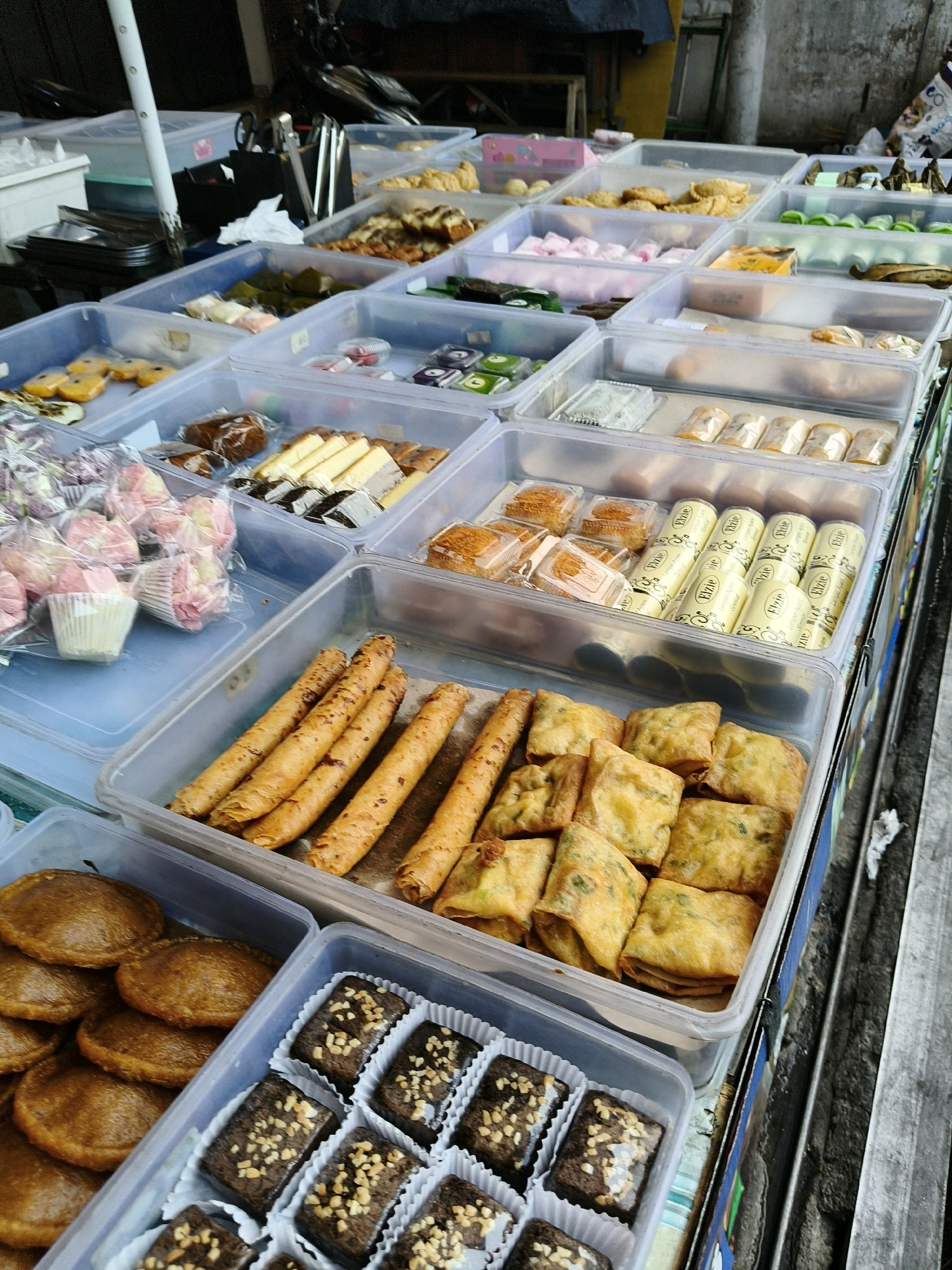 The image depicts an outdoor food stall in Bandung City, Indonesia, displaying a variety of Indonesian snacks and pastries. The stall features multiple clear plastic containers filled with different food items arranged in rows. In the foreground, a container holds round, flat, golden-brown cakes, possibly *kue apem*. Next to it, another container showcases dark brown, square brownies topped with chopped nuts, individually placed in white paper liners. Behind these, a container is filled with golden-brown, rectangular savory snacks, likely *martabak telur*. To the left, a container displays long, cylindrical, golden-brown crispy snacks, possibly *semprong* or *kue gabus*. Further back, other containers hold various sweets, including pink meringue-like cookies, small cakes, and green-wrapped items that could be *kue lapis*. The stall is set up on a street with a concrete wall visible in the background and a portion of a motorcycle parked behind the stall. The overall scene is bright and suggests a bustling marketplace environment.