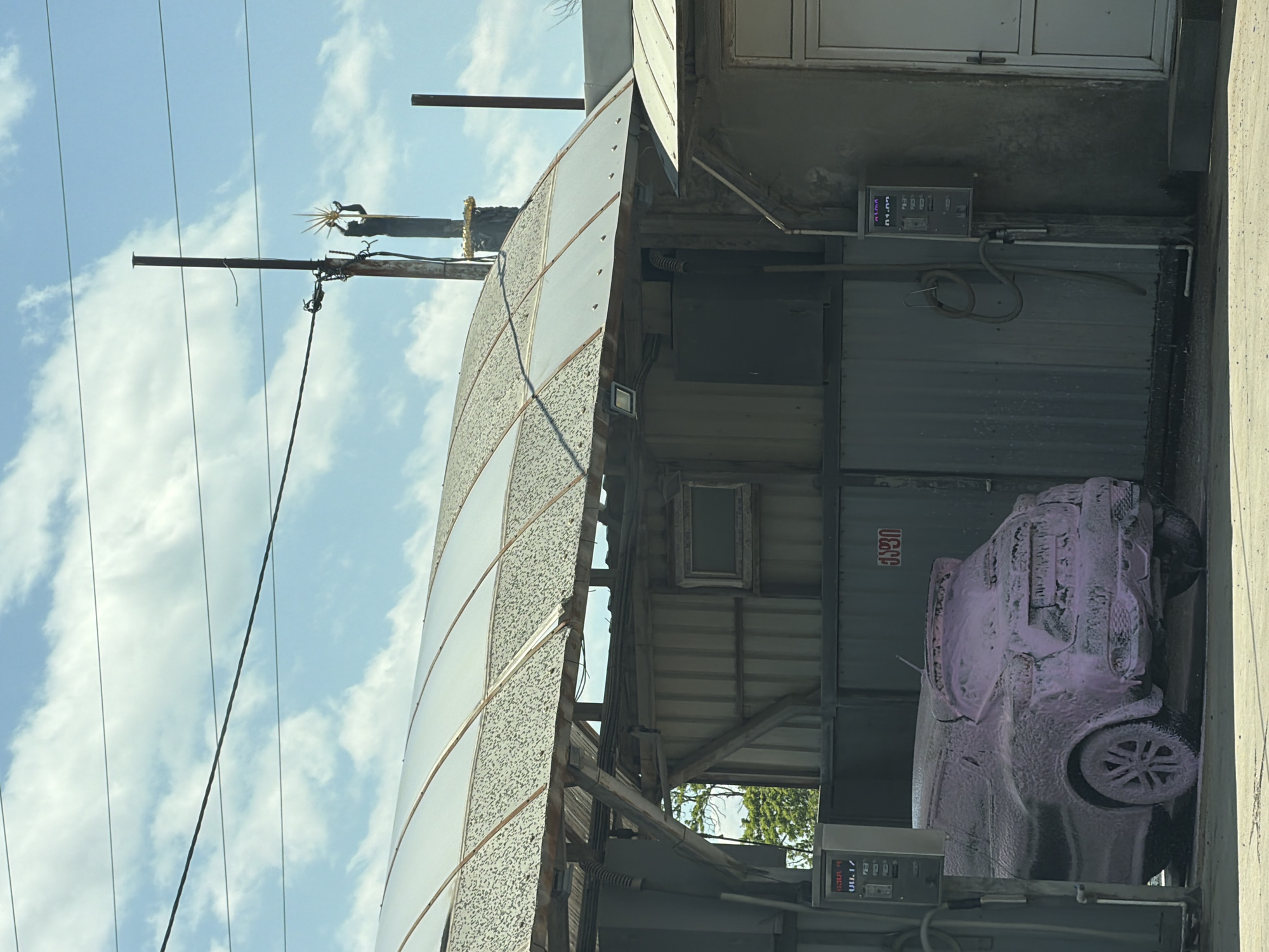 The image shows a car being washed in a car wash in Tbilisi, Georgia. The car is covered in pink foam. The car wash appears to be an older, utilitarian structure. Power lines are visible in the background against a partly cloudy sky.