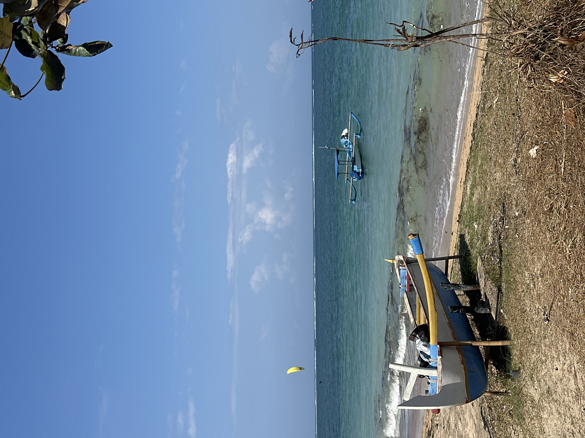 An eye-level, medium shot captures a serene beach scene in Denpasar, Indonesia, under a clear blue sky dotted with scattered white clouds. In the foreground, a traditional Balinese outrigger boat, painted white with blue and yellow accents, rests on wooden supports on the sandy shore. The boat appears empty except for some dark bags inside. The sand is covered with dry grass and some scattered debris.
The turquoise water of the ocean gently laps at the shore, with small waves breaking. In the mid-ground, another blue and white outrigger boat with a canopy is anchored in the water. Further out in the ocean, a solitary kite surfer is visible, their yellow kite a bright speck against the horizon. The horizon line is marked by a thin white line, likely the edge of a reef or distant land. The scene is framed on the right by the dry, spiky branches of a plant. The overall atmosphere is one of tranquility and natural beauty.