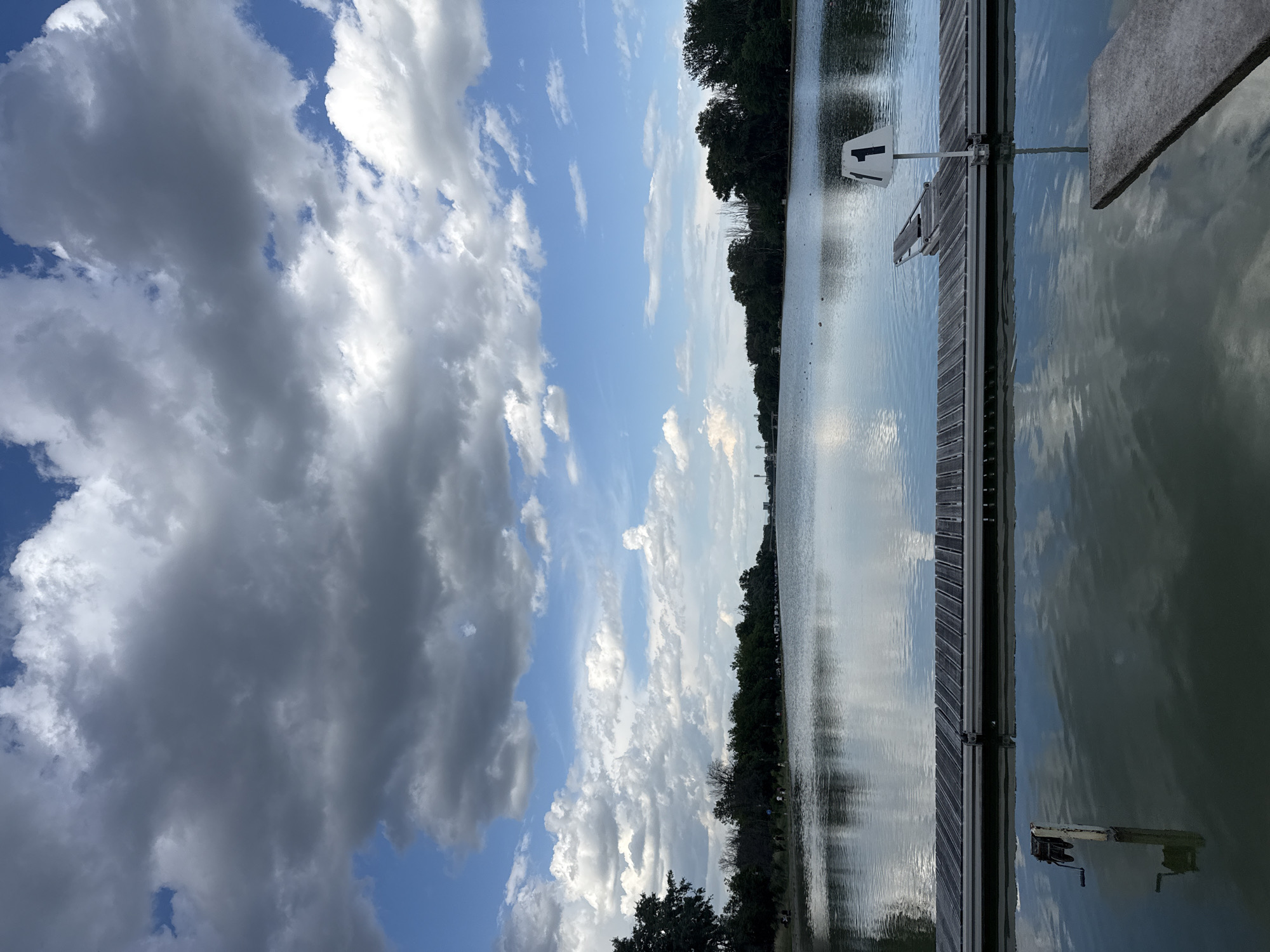 The image shows a section of the Rowing Canal in Plovdiv, Bulgaria. The view is from the water's edge, looking towards the canal. A wooden dock is visible on the right side of the frame, with a sign displaying the number '1'. The canal itself is calm, with reflections of the sky and clouds. The sky is partly cloudy, with dark, heavy clouds on the left and patches of blue sky. A line of trees is present along the opposite bank of the canal. The orientation of the image is rotated 90 degrees counterclockwise.