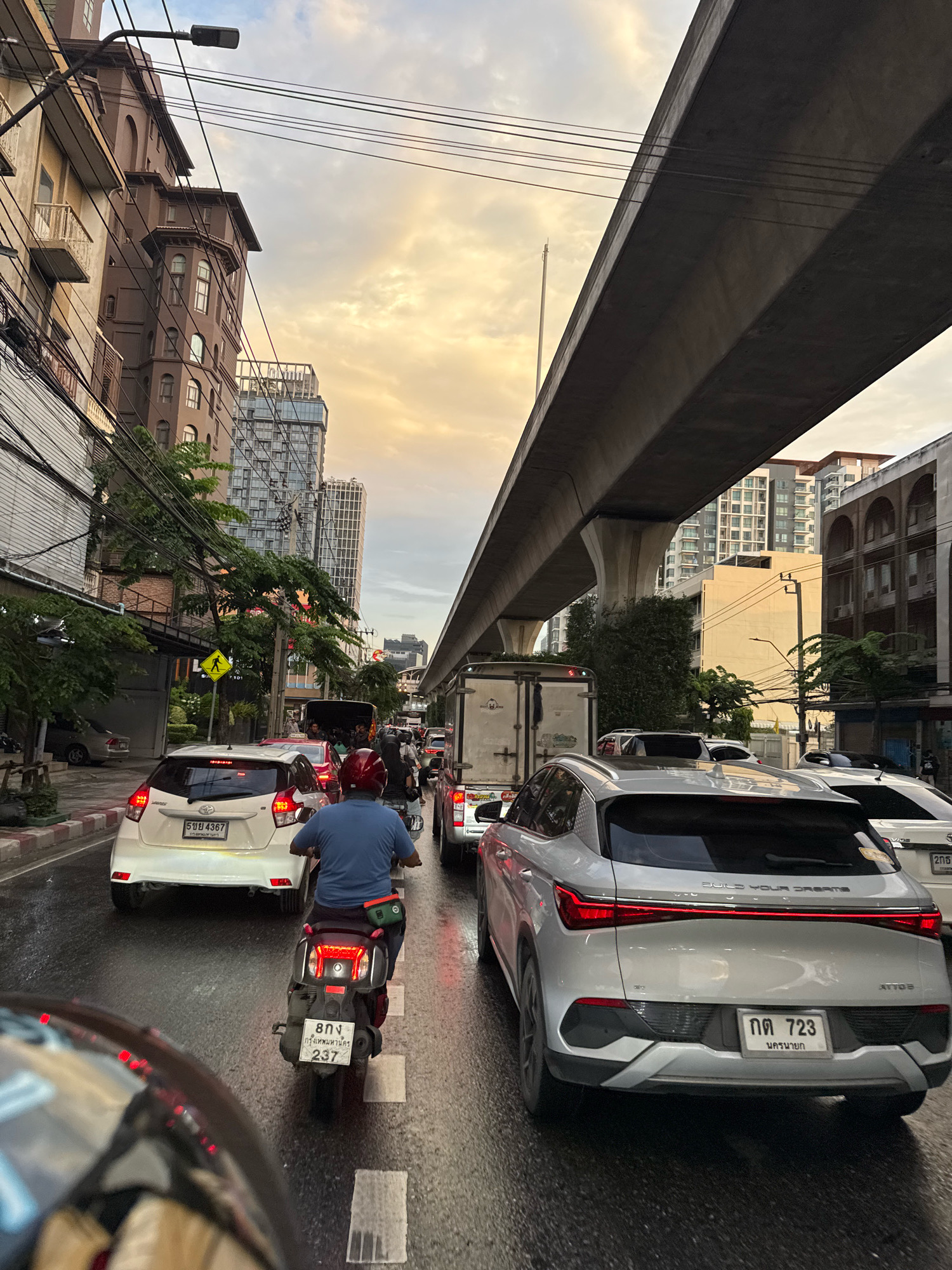 This is a ground-level, eye-level, full shot of a traffic jam on a city street in Bangkok, Thailand, likely during sunset or overcast conditions due to the lighting and sky color. The road is wet, indicating recent rain.
In the foreground, a motorcycle with a rider wearing a blue shirt and a red helmet is visible, stopped in traffic. The motorcycle has a license plate that reads "808 กรุงเทพมหานคร 237". To the right of the motorcycle, the rear of a silver SUV is prominent. This SUV has a distinctive LED taillight bar and the words "BUILD YOUR DREAMS" inscribed on its rear. Its license plate reads "กก 723 นครมงคล".
Further ahead in the traffic lane, a white hatchback is seen, with its license plate partially visible as "508 4367". Beyond this, a large white box truck and other vehicles are part of the congestion. Overhead, a concrete elevated structure, likely part of an expressway or sky train, casts a shadow over the scene.
The background features several multi-story buildings, including a dark brown building with architectural details and balconies on the left, and taller modern buildings with many windows in the distance. Trees line the street, and electrical wires crisscross above. A yellow pedestrian crossing sign is visible on the left side of the road. The overall impression is one of a busy urban environment experiencing significant traffic congestion.