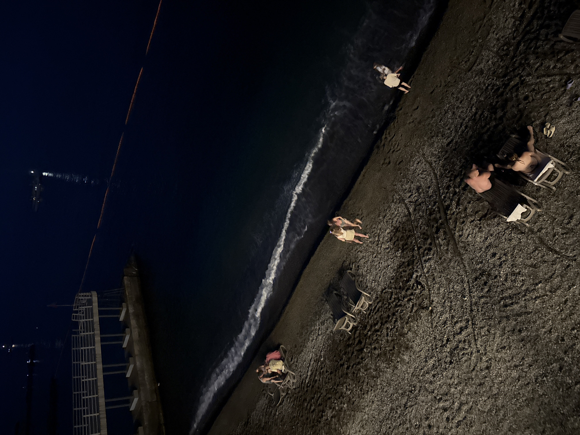 At nighttime, on a pebbled beach in Yalta, Ukraine, several people are visible. On the left side of the image, four individuals are gathered around a lounger, appearing to be engaged in conversation. Further down the beach, two people are standing and walking. Near the water's edge, two more individuals are standing and looking out at the sea. In the foreground, two more people are seated on loungers, with one appearing to be relaxing and the other possibly receiving a massage. The dark sea is visible with gentle waves breaking on the shore. In the background, a pier with railings extends into the water, and a boat with lights is visible in the distance. The overall scene suggests a relaxed evening atmosphere on the beach.