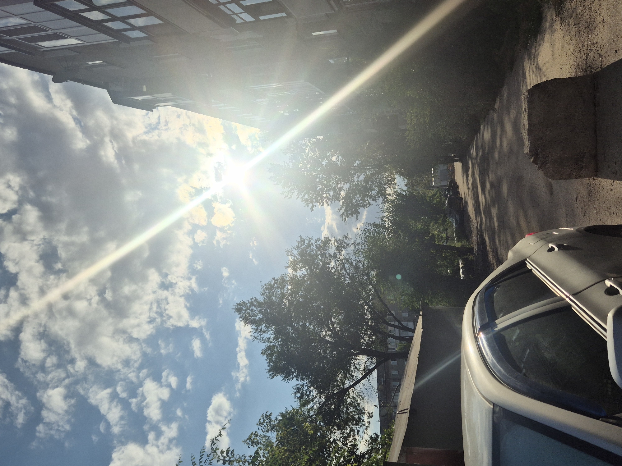 The image shows a sunny day in Kokshetau, Kazakhstan, with a car parked on a dirt road next to a building. The sun is bright and casts long shadows. The car is white and appears to be a sedan. The building is a multi-story apartment building. Trees line the road, and there are some clouds in the sky. In the foreground, there is a large concrete block on the side of the road. The scene appears to be in a residential area.