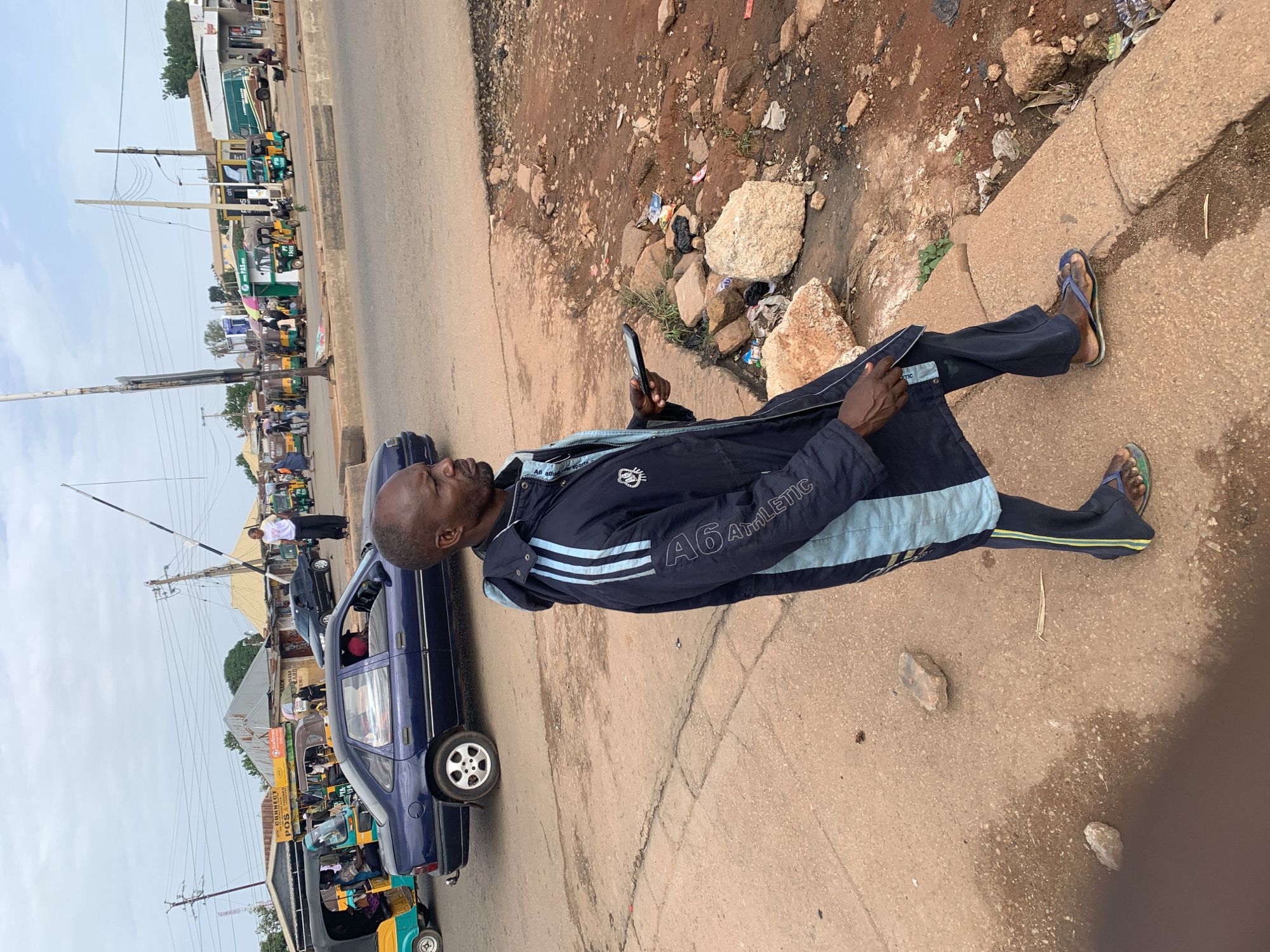 An African man stands on a dirt and concrete embankment, looking to his right. He wears a dark blue jacket with light blue stripes down the sleeves and light blue accents on the sides of the pants. He holds a black smartphone in his right hand. The man is positioned in the foreground, with a busy street scene in the background. In the background, there are several vehicles, including a dark blue sedan, multiple yellow and green auto-rickshaws, and a white van. Shops and buildings line the street. The sky is overcast. The scene appears to be a street corner in Gura Top, Nigeria.
