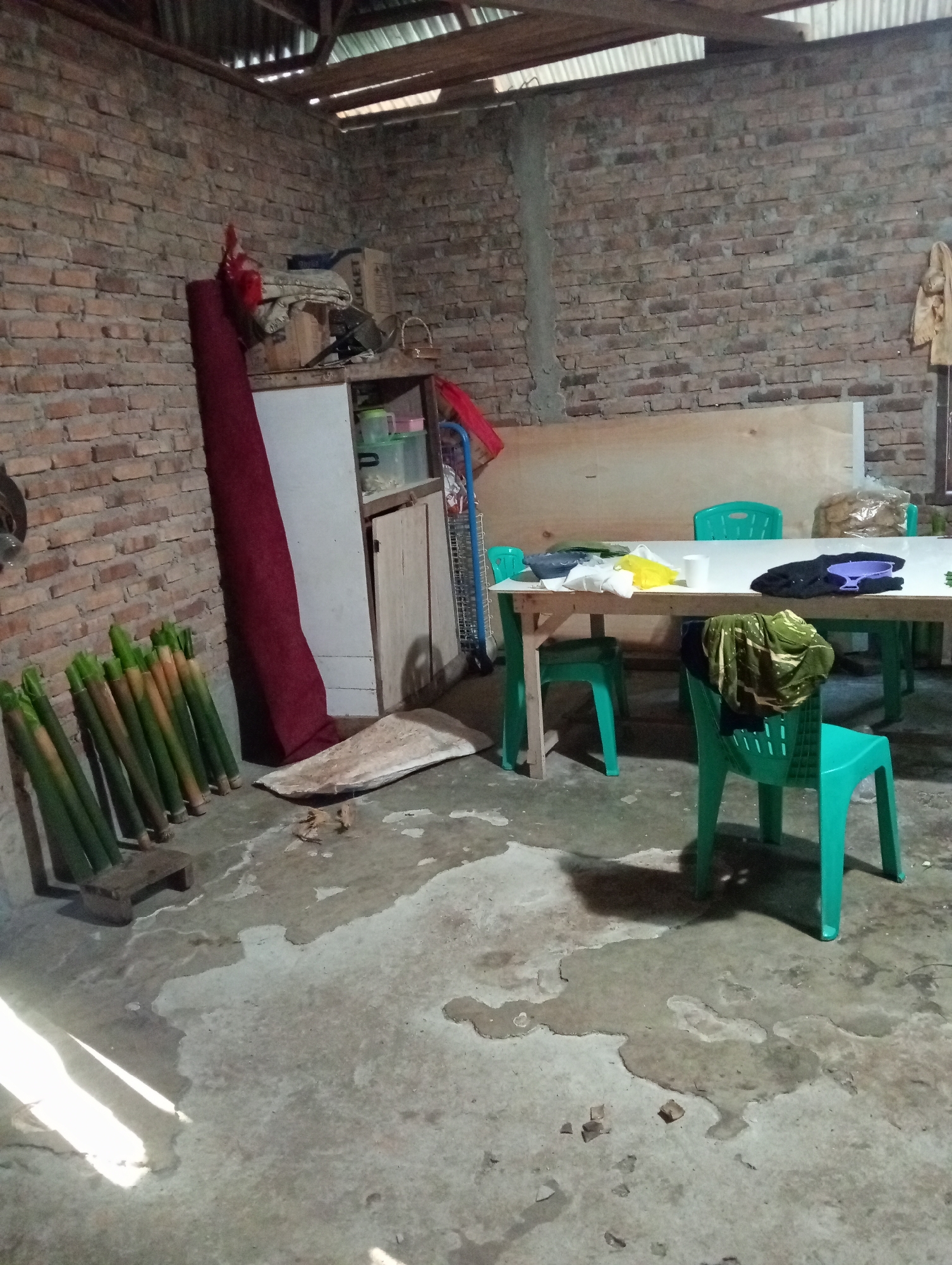 This image depicts an indoor scene, likely a room in a residence or a small workshop, in Campago Barat, Indonesia. The room has unfinished brick walls and a corrugated metal roof with visible wooden beams. The floor is made of concrete and shows signs of wear and some water stains.
On the left side of the image, a stack of long, green bamboo-like stalks is leaning against the wall, with a small wooden object at their base. To the right of the stalks, a large roll of dark red fabric stands upright. Adjacent to the fabric is a white cabinet with a wooden door, and on top of it are several containers and boxes. Further to the right, a long wooden table is set up with several bright green plastic chairs around it. On the table are various items including a white cup, a yellow cloth, a bowl, and some bagged items that appear to be snacks or food. A dark piece of clothing with a purple graphic is draped over the table, and another green and brown patterned cloth is draped over the back of one of the chairs. The overall impression is one of a functional, possibly domestic or small-scale production space.