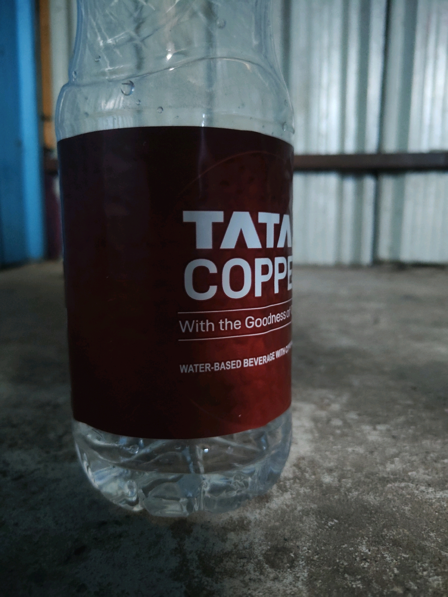 A partially filled, clear plastic bottle of Tata Copper water rests on a textured, concrete surface. The bottle has a maroon label with white text that reads "TATA COPPER" and "With the Goodness of" and "WATER-BASED BEVERAGE WITH CO". The background shows vertical corrugated metal sheeting, possibly part of a structure or fence, and a blurred blue vertical element. The lighting is dim, creating shadows on the concrete. There are no people present, and no action is depicted. The scene appears to be outdoors in an urban or semi-urban setting, possibly in Endada, India, given the context provided.