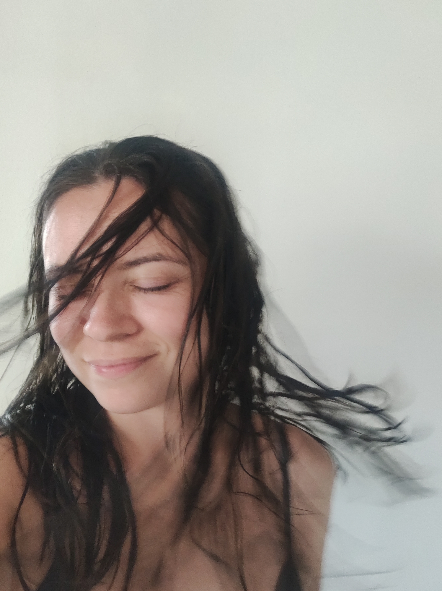 A female subject is depicted in a medium-close-up shot against an unidentifiable light-colored background. The subject has long, dark brown hair, which appears wet and is actively moving, with multiple strands obscuring portions of her face, particularly her left eye and forehead. Her eyes are closed, and her lips are upturned in a gentle smile. The visible skin tone is light-to-medium. Portions of her bare shoulders and upper chest are visible, suggesting minimal clothing or a garment with thin straps. The background consists of a smooth, uniform light gray or off-white surface, devoid of any distinguishing features or objects. No specific geographic location or city, including Фоки, Russia, can be determined from the visual content of the image.