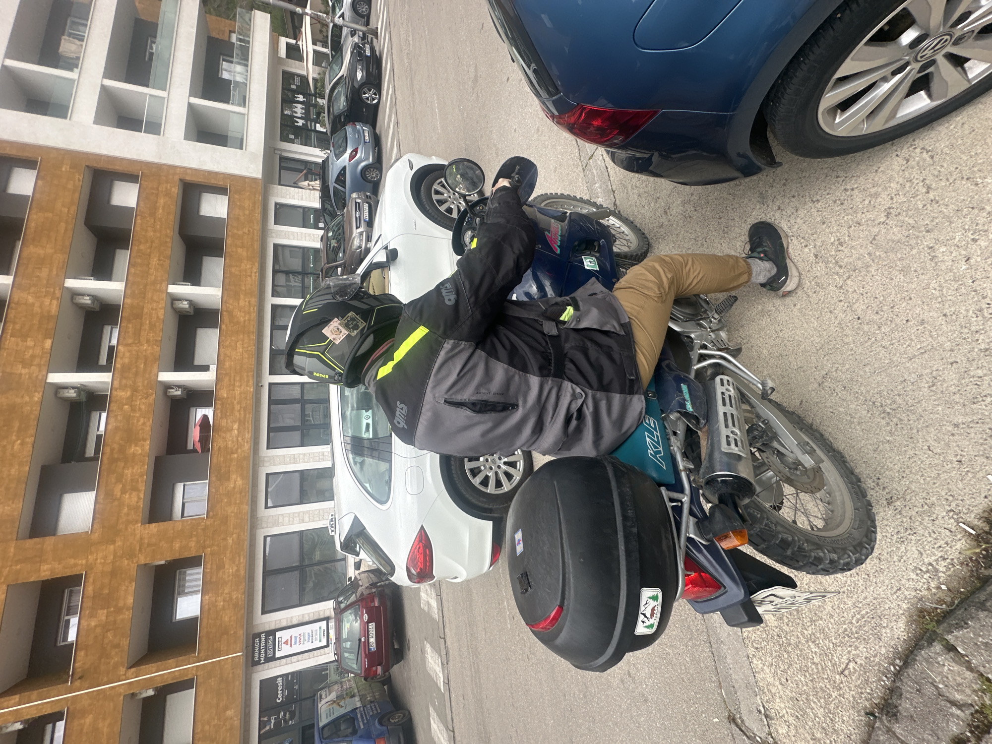 A man wearing a motorcycle helmet and jacket sits on a blue motorcycle in Budva, Montenegro. The bike is parked on a street, alongside other parked cars, including a blue Volkswagen to the right and a white car to the left. The building to the left is partially visible. It has a facade with a mixture of light brown and white architectural elements. In the background, a business sign is visible on a building behind the white car. The motorcycle has a black storage box attached to the back, and the license plate is visible.
