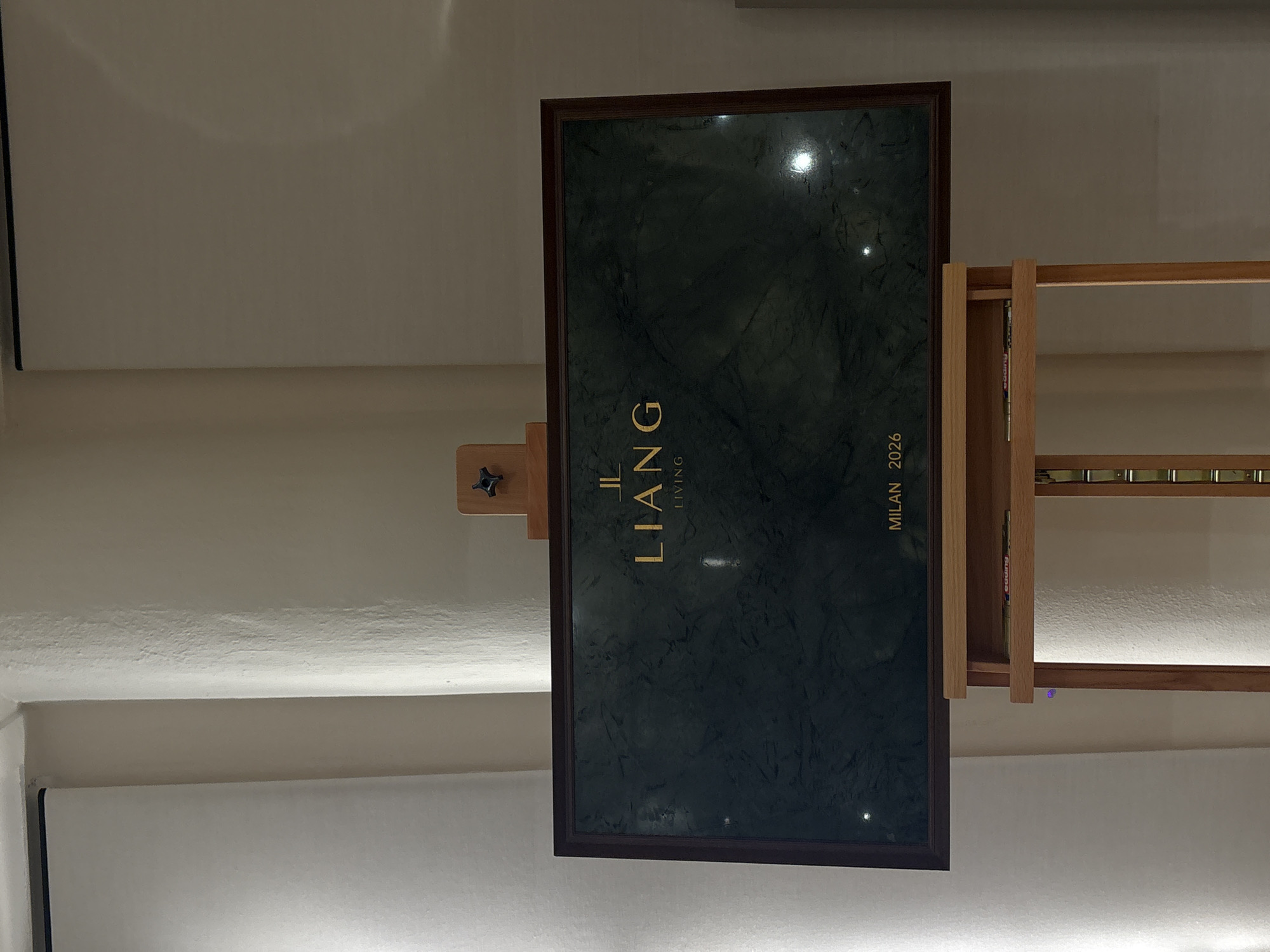 The image displays an indoor setting featuring a vertically oriented, framed panel, likely part of an exhibition or showroom. The main subject is a large, rectangular panel with a dark green, marble-like surface, encased in a dark wooden frame. Prominent gold lettering is visible on the panel: "LIANG LIVING" runs vertically up the left side, and "MILAN 2026" runs vertically up the right side.
The panel is supported by a light-colored wooden easel or stand, which has a visible black adjustment knob. To the right of the framed panel, a light wooden shelving unit or display rack is partially visible, containing several small, cylindrical metallic objects on one of its shelves.
The background consists of plain, light-colored walls, possibly white or off-white. A horizontal line of strip lighting illuminates the lower section of the wall, creating a soft glow. Reflections from overhead or unseen light sources are apparent on the polished surface of the dark green panel. The overall aesthetic is clean and modern, suggesting a presentation of a brand or collection. The visible text "MILAN 2026" indicates a future event, launch, or collection associated with the city of Milan, Italy. No people are visible in the scene.