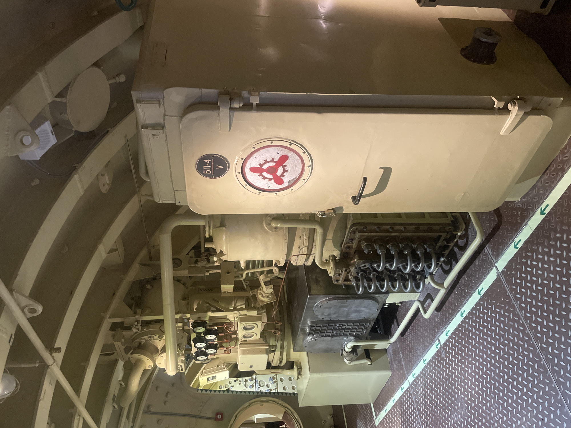 This image shows the interior of a Soviet-era submarine at the Kronstadt Naval Museum in Kronstadt, Russia. The scene depicts a section of the submarine's engine room or a technical compartment, characterized by complex machinery, pipes, valves, and gauges. The walls and ceiling are painted a muted beige or olive green, typical of military vessels.
Dominating the right side of the frame is a large, cream-colored cabinet or hatchway with a circular emblem featuring a red propeller-like symbol against a white background, and above it, a Cyrillic inscription that reads "БП-4 V". A black handle is visible on the front of this unit.
To the left, a dense arrangement of equipment includes what appears to be a heat exchanger or radiator with a series of coiled tubes, connected to a larger metal box. Several gauges with black faces and white markings are mounted on a panel above other machinery. Copper piping is visible throughout the machinery, indicating a complex system of fluid or gas transport.
The floor is covered with diamond-plate metal flooring, commonly found in industrial and maritime settings, with green directional arrows painted on it, suggesting a pathway. The overall lighting is subdued, with some areas illuminated by overhead light fixtures, casting shadows and highlighting the textures of the metal surfaces. No people are present in the image.