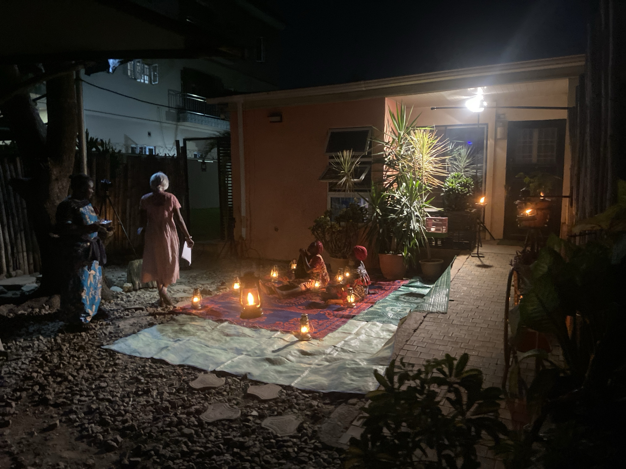 At night in Barden Yana, Nigeria, a gathering takes place in a backyard. Two individuals stand to the left on a gravel surface, one clad in a dress and the other in traditional attire, facing the center. A rectangular tarp covers a portion of the gravel, extending towards a brick pathway. On the tarp, a colored rug is illuminated by several oil lanterns, with figures seated on the rug. The backyard is bordered by a single-story building with a porch light and visible plants. The backdrop includes a building with windows and a balcony.