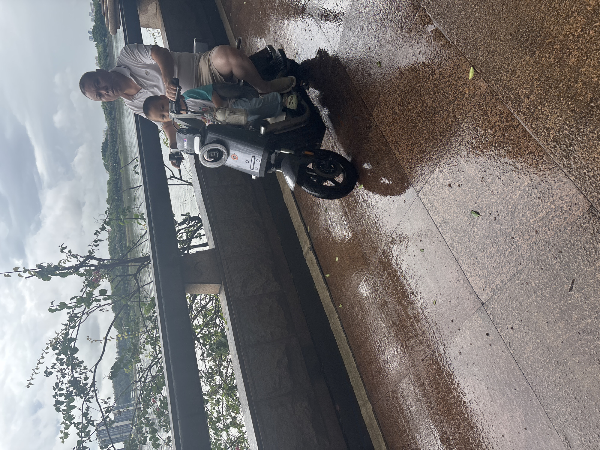 A man and a young boy are riding an electric scooter along a wet, tiled pathway. The man is driving, wearing a white polo shirt and light-colored shorts, while the boy sits in front of him. The boy is wearing a t-shirt with blue and white sleeves and light blue pants. The scooter is dark gray with a prominent circular headlight and a bag hanging from the handlebars. The pathway appears to be made of large, textured brown tiles that are wet, reflecting the overcast sky and surrounding environment. In the background, a dark railing separates the pathway from a body of water, beyond which are trees and buildings. The scene suggests an outdoor location, possibly a park or waterfront area in Nanhai District, China, given the overcast weather and the presence of typical urban elements.
