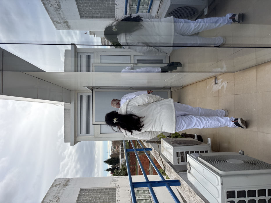The scene captures three individuals walking along an outdoor balcony corridor on an upper floor of a building in Meknes, Morocco, under a cloudy sky. The foreground subject, viewed from behind, has long dark hair adorned with a white flower or clip, and is wearing a white fluffy jacket, white trousers, and dark athletic shoes with white soles and red accents. This individual walks towards an open doorway. Ahead, two additional individuals, both wearing white attire consistent with medical or professional uniforms, also proceed towards the doorway. To the left, a blue metal railing borders the balcony, with two white outdoor air conditioning units mounted along the wall below it. Beyond the railing, a view of trees and distant buildings is visible under the overcast sky. A large vertical glass panel on the right reflects the foreground subject and the immediate environment.