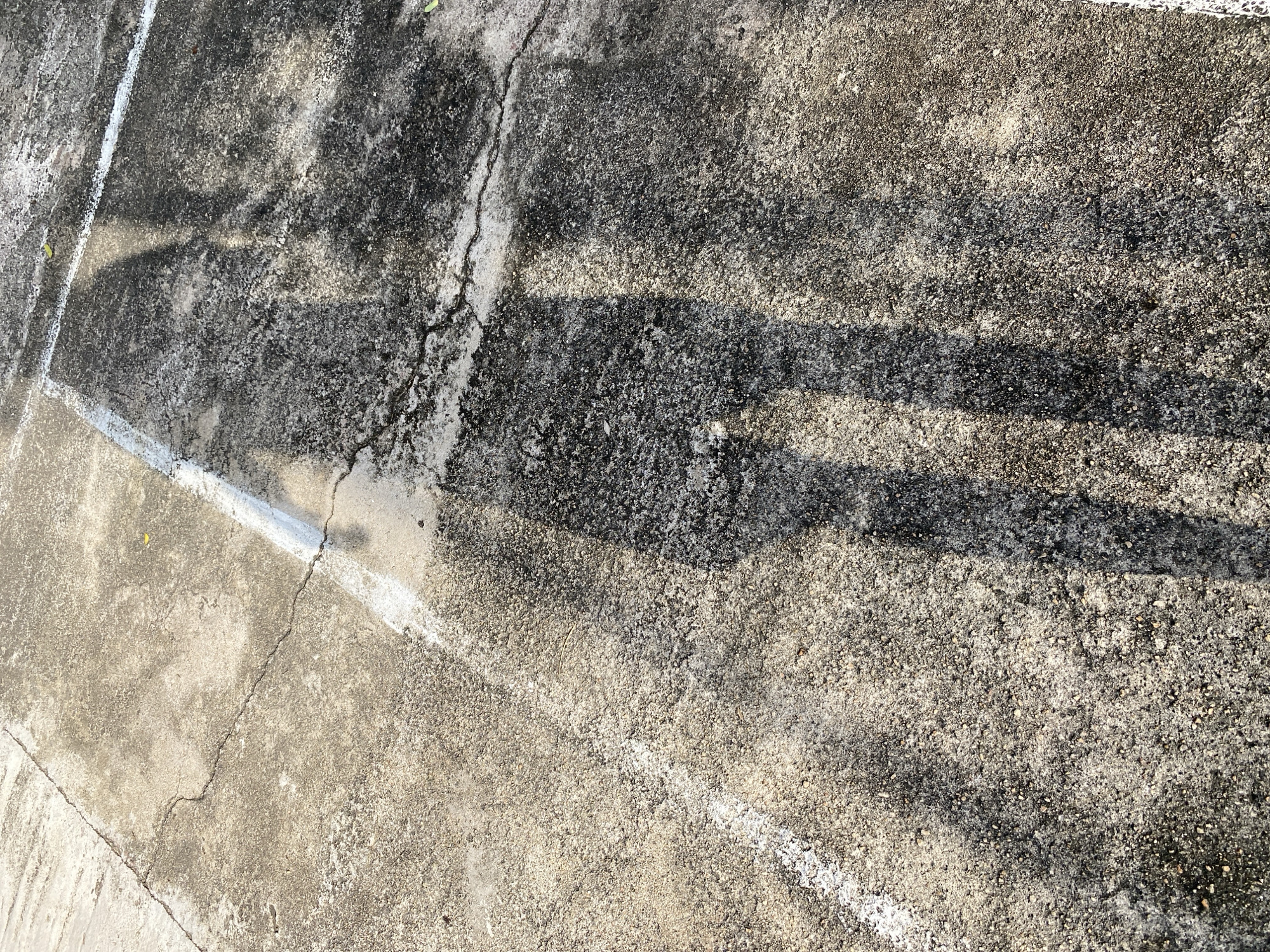 The image displays a shadow of a person on a cracked, weathered concrete surface. White lines, possibly markings from a court or boundary, are visible on the concrete. The lighting suggests it is daytime, with sunlight casting the shadow. The environment appears to be outdoors, likely in a public space. The scene is devoid of any explicit human presence, with only the shadow indicating a person's form. The texture of the concrete is rough and granular, with areas of discoloration and signs of age. A small green leaf is present on the concrete in the upper right portion of the image. Without additional information, it is not possible to definitively place this image in Jalingo, Nigeria.