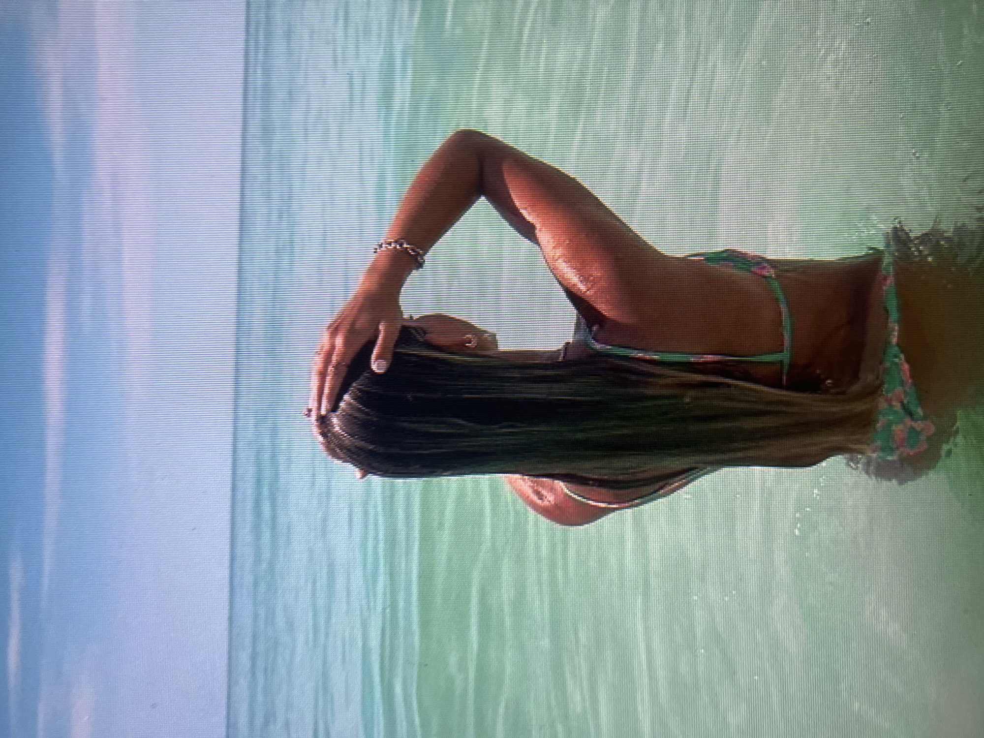A woman stands in clear, turquoise water, with her back to the camera. She is wearing a patterned bikini. Her hands are raised, one resting on her head and the other behind her head. Her long, dark hair flows down her back. The ocean stretches out before her, meeting a pale blue sky with wispy clouds. The image is captured from the chest up.