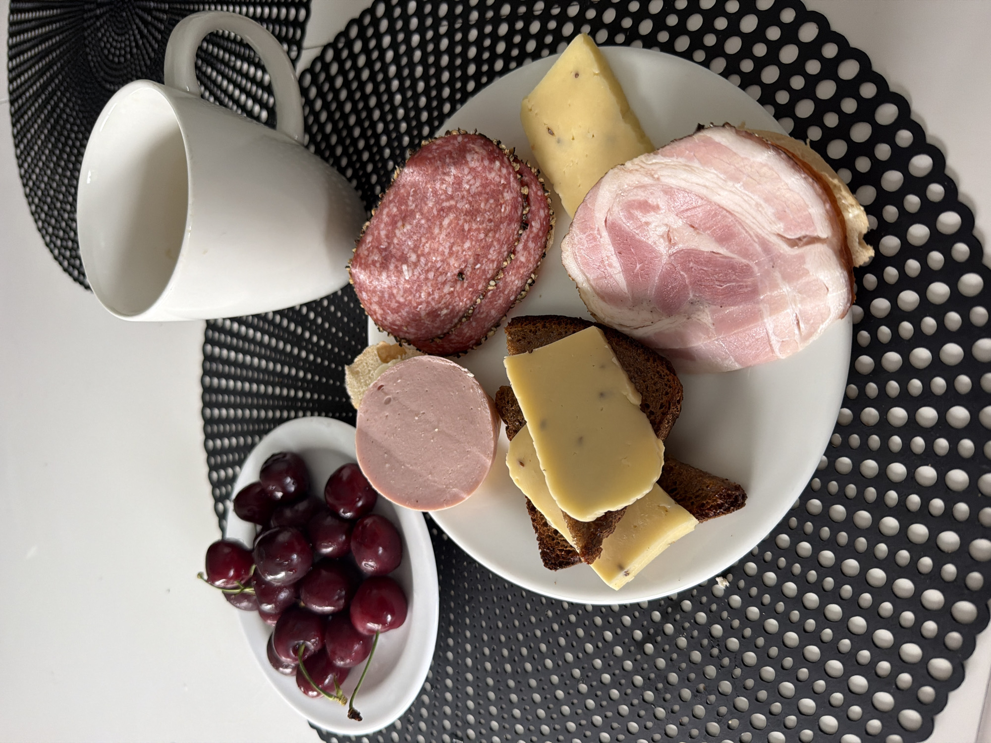 A plate of food sits on a black placemat on a white table. The plate contains several slices of cheese on pieces of dark bread, two slices of salami, a round slice of ham, and a slice of cured bacon. To the left of the plate, on a smaller white plate, is a pile of dark red cherries. A white mug sits to the right of the plate. The image was taken in Baloži, Latvia.