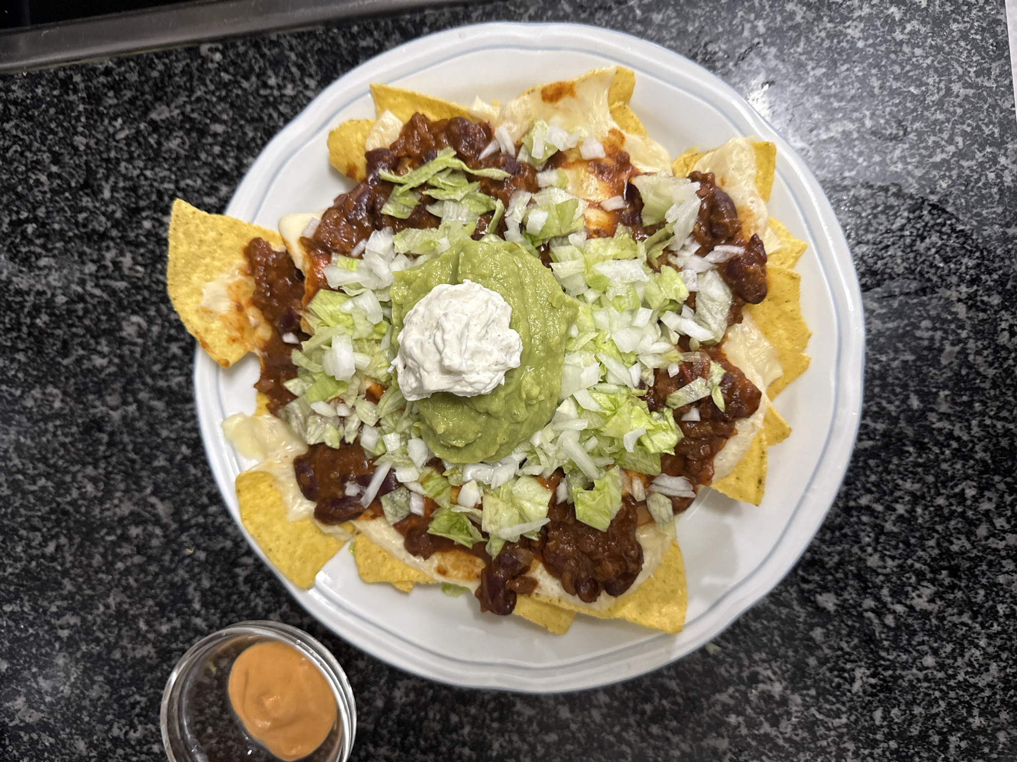 The image shows a plate of nachos on a dark, speckled countertop. The nachos consist of tortilla chips topped with melted cheese, chili, shredded lettuce, guacamole, and sour cream. A small dish of sauce is next to the plate. The location is not discernible from the image.
