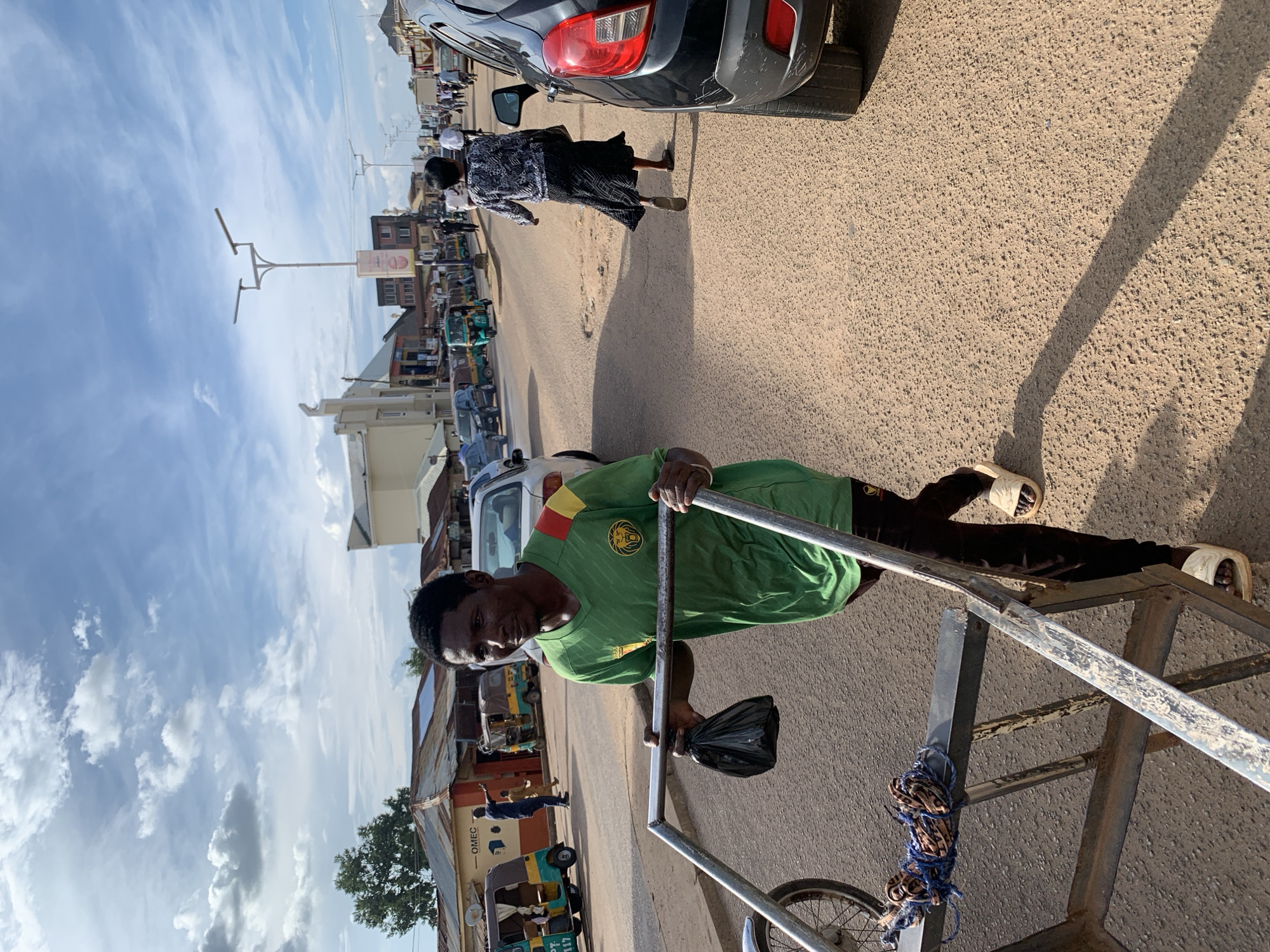 In Jos, Nigeria, under a bright sky with scattered clouds, a man in a green t-shirt and black pants pushes a metal handcart on a dusty, paved road. The man, with dark skin and short black hair, is looking to his right and appears to be mid-stride, his left hand gripping the cart's handle and his right hand holding a black plastic bag. The handcart, made of silver metal, has a wheel on the left side and a bundle of rope tied to its platform.
The road is lined with buildings on the left, including some with corrugated metal roofs and one with a sign reading "OMEC." Several colorful auto-rickshaws are parked along the side of the road, along with other vehicles. On the right side of the road, a black car is partially visible, with its rear bumper and taillight showing. Further down the road, other pedestrians and vehicles are present, suggesting a typical urban scene. Streetlights with multiple arms are also visible, indicating street lighting infrastructure. The overall impression is a bustling street scene in a Nigerian city.