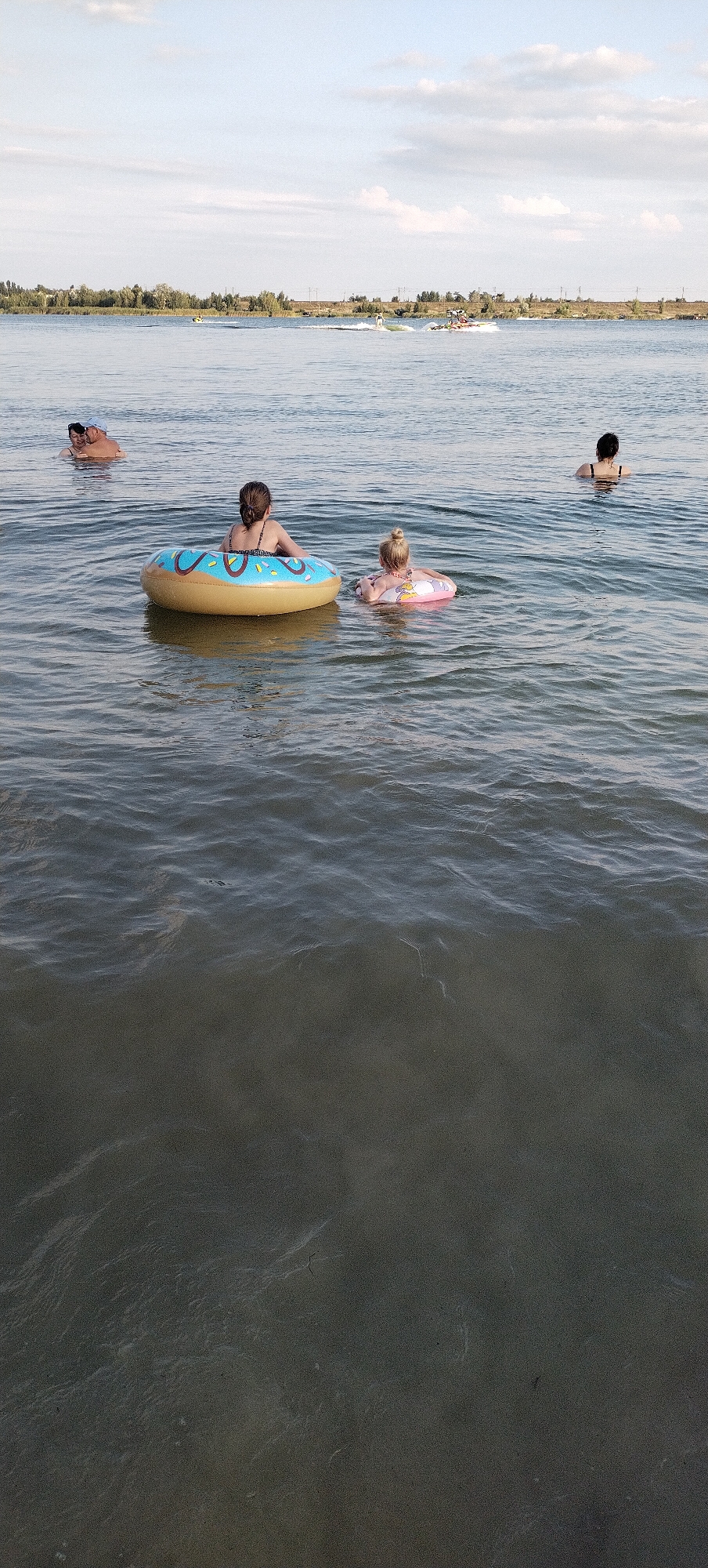 An outdoor, medium-distance, eye-level shot captures a group of people enjoying a body of water. The scene appears to be on a lake or river, with a distant shoreline featuring trees and structures. The water is a murky green, with visible ripples and waves. In the foreground, two young girls are present. One girl, with blonde hair tied in a bun, is floating on a pink inner tube. Next to her, a girl with dark hair, wearing a patterned swimsuit, sits in a large, yellow inflatable donut. Further back, two adults, appearing to be a man and woman, are partially submerged in the water, embracing. The background shows several individuals on jet skis, creating wakes in the water, and a few distant boats. The sky is a pale blue with scattered clouds. The location is identified as Bezliudivka, Ukraine.
